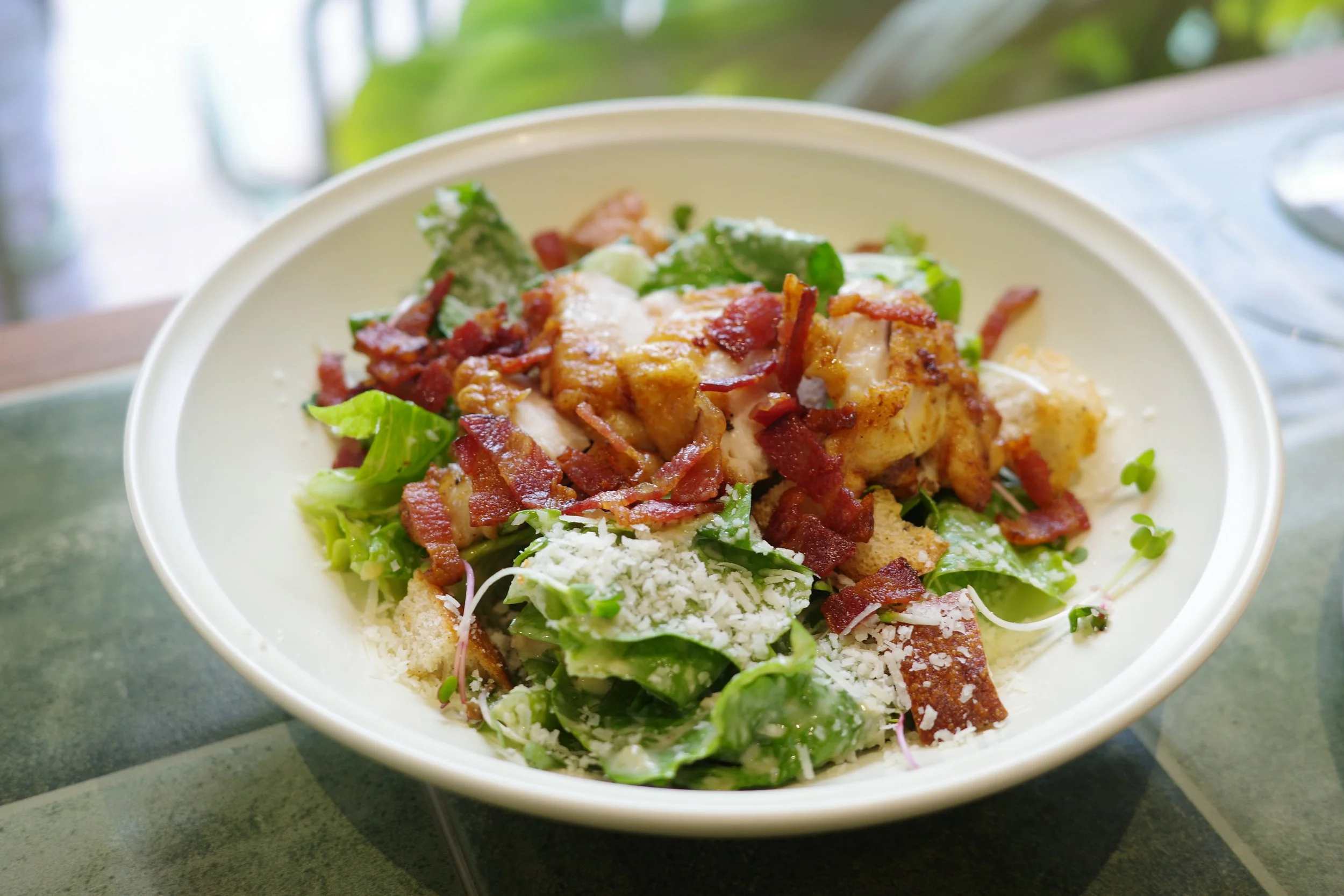 Bowl of Caesar salad with romaine lettuce, bacon bits, shredded cheese, croutons, and grilled chicken.