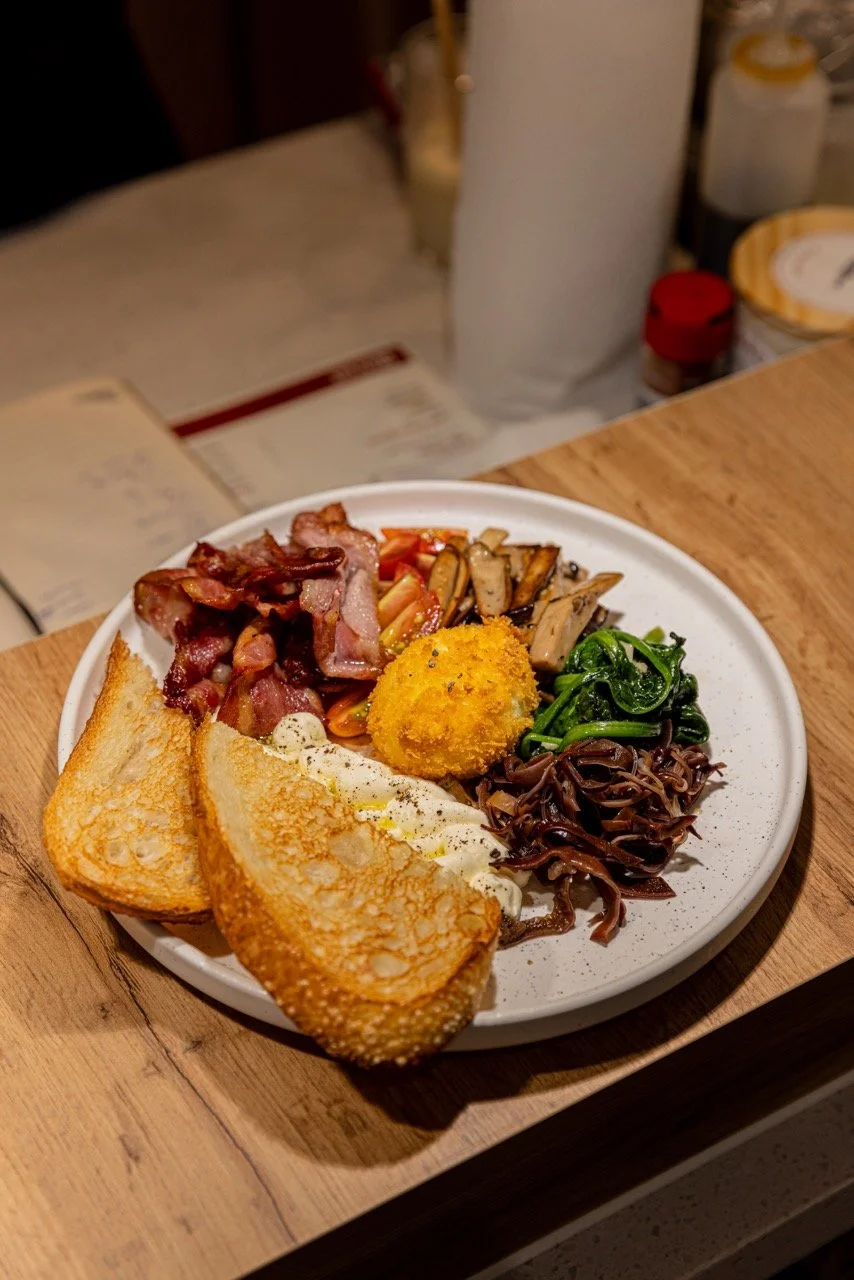 A plate of breakfast foods including bacon, sautéed mushrooms, sautéed spinach, fried egg, hash brown, and toasted bread slices.