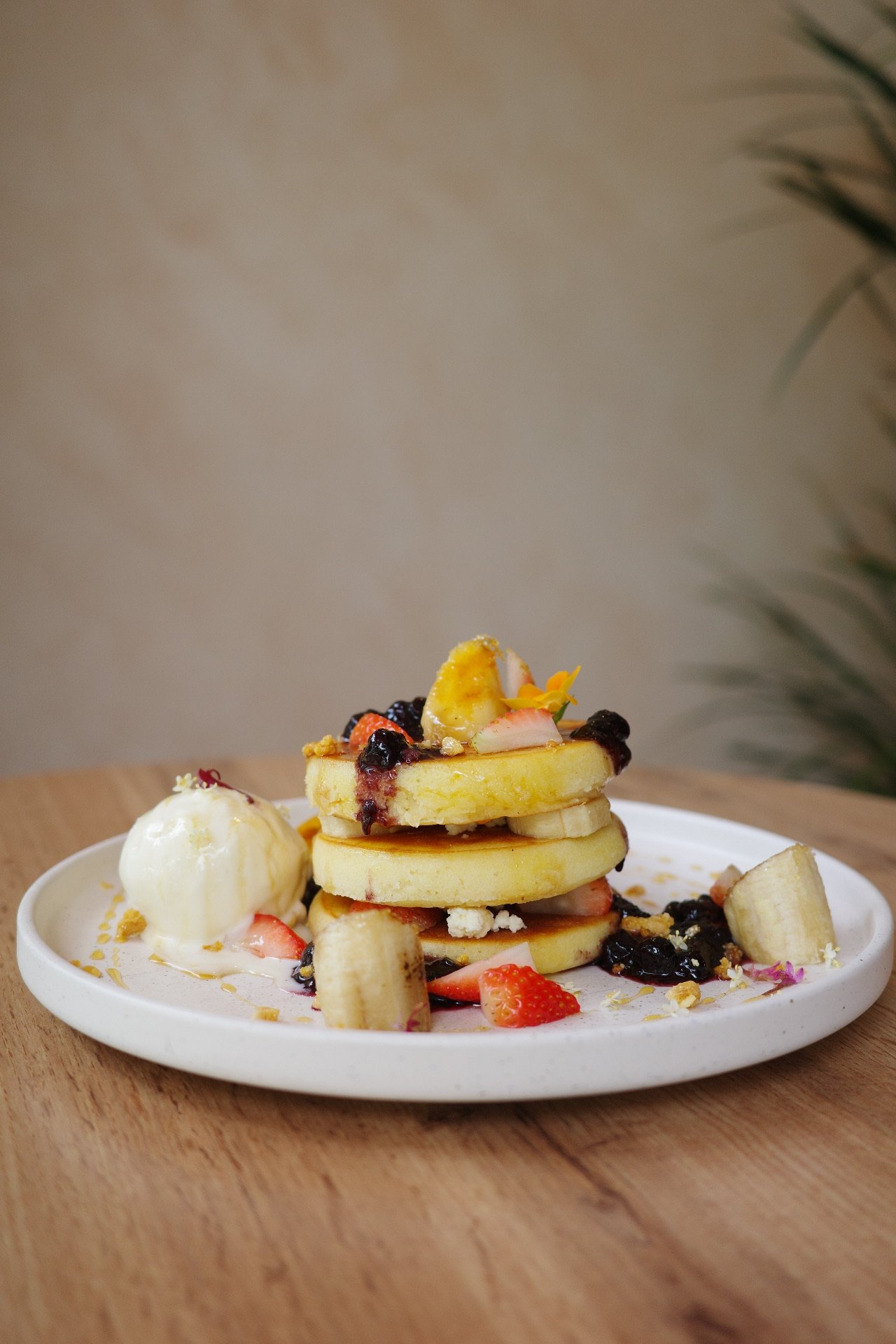 Stacked pancakes topped with sliced bananas, strawberries, blueberries, and edible flowers, served with a scoop of ice cream on a white plate.