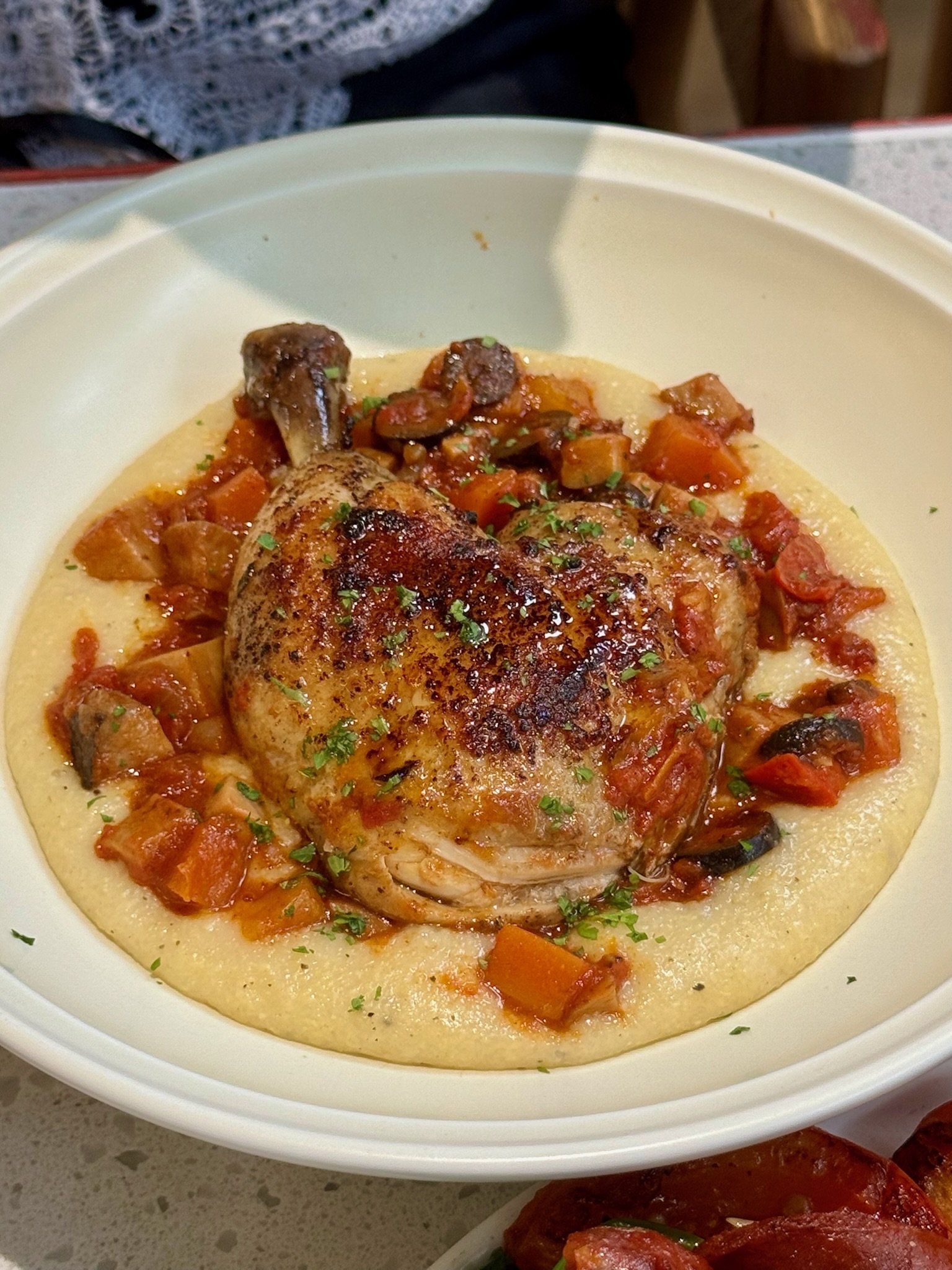 A white bowl with cooked chicken on a bed of creamy sauce, topped with chopped parsley, surrounded by a vegetable ragout with diced carrots, tomatoes, and mushrooms.