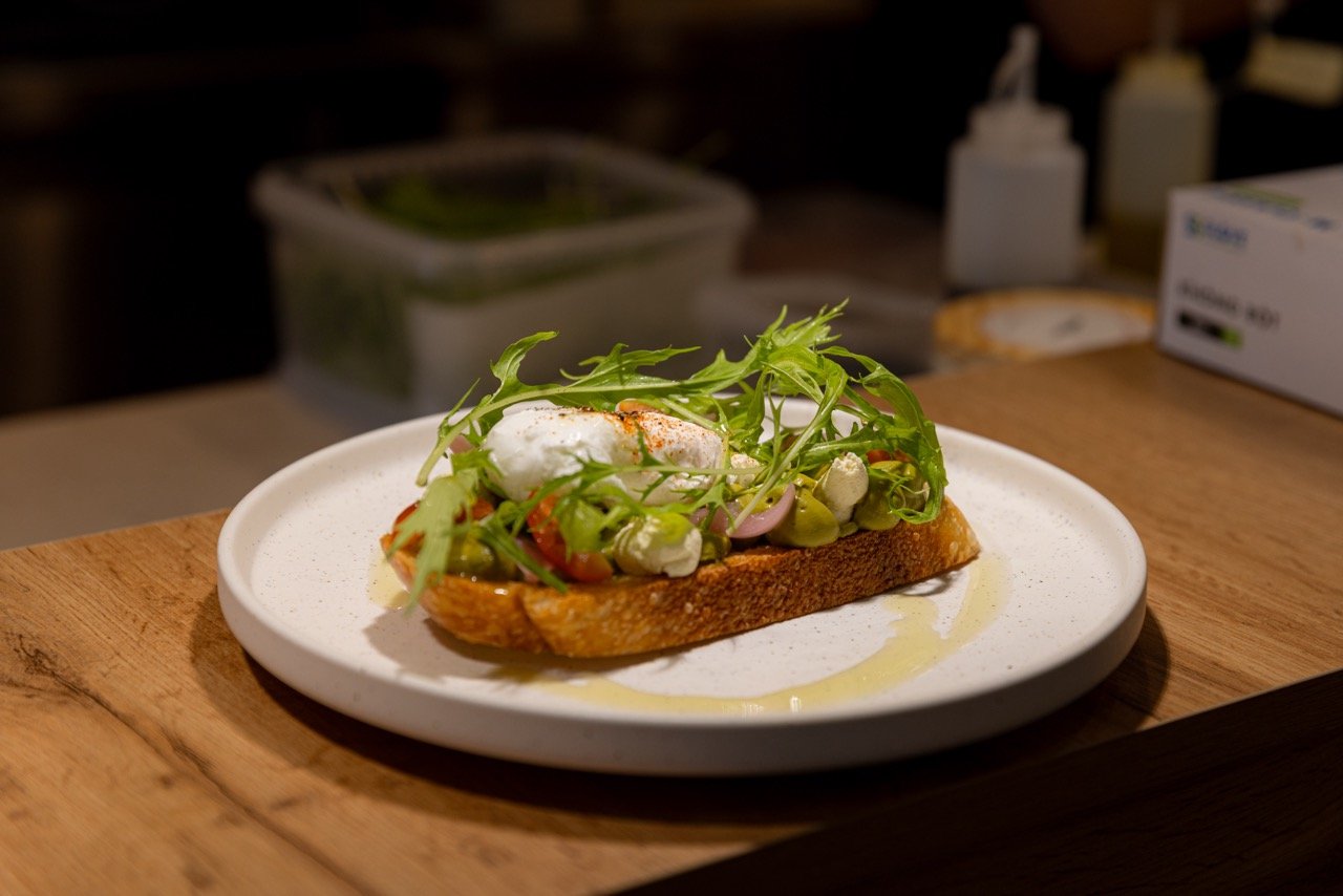 Open-faced sandwich on a white plate with toasted bread, topped with greens, vegetables, and a dollop of sauce or cheese.