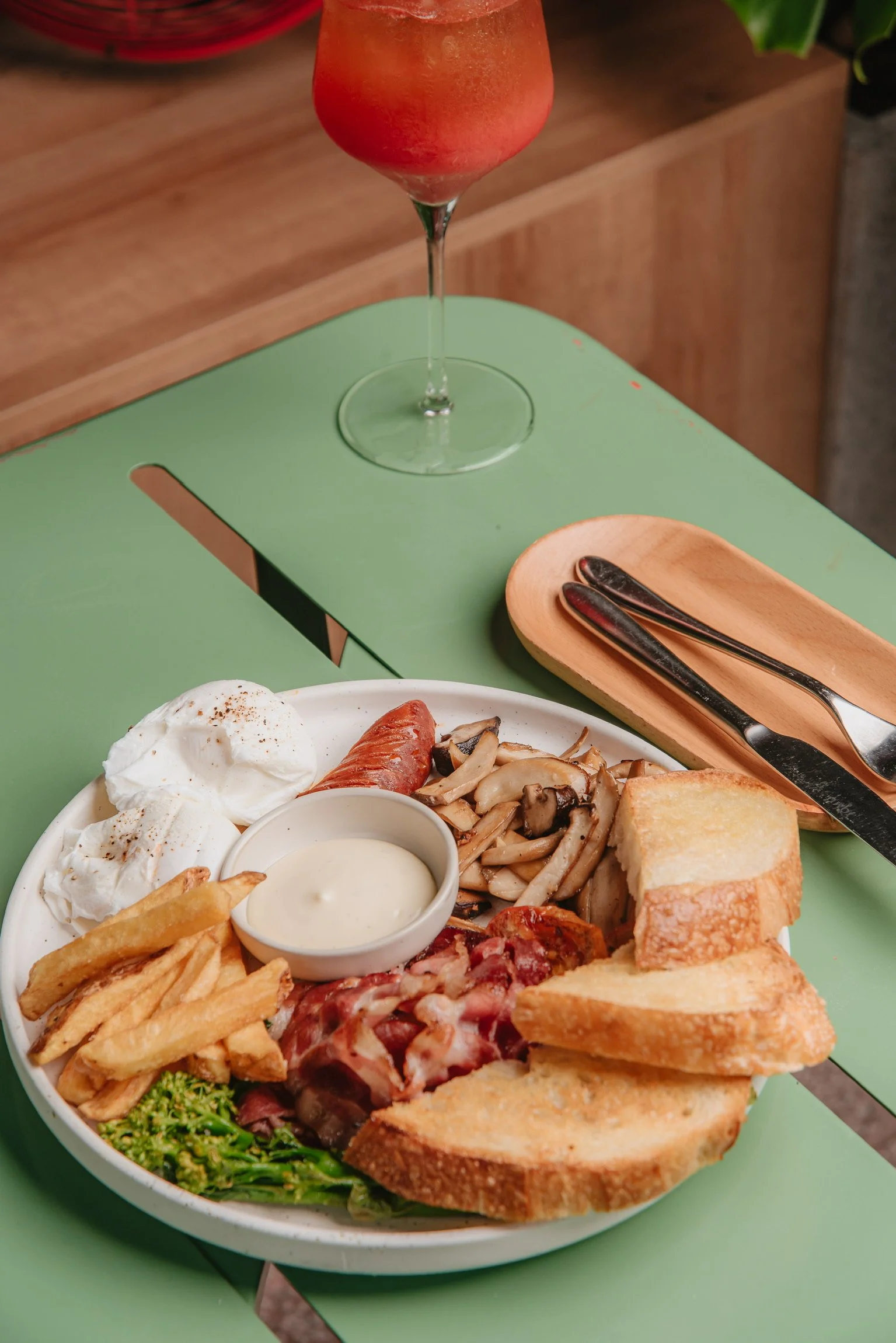Close-up of a large plate of assorted breakfast foods, including eggs, French fries, toast, bacon, sausage, and broccoli, with a small bowl of dipping sauce, set on a green table. In the background, a tall glass of orange-pink mixed drink on a green tabletop.