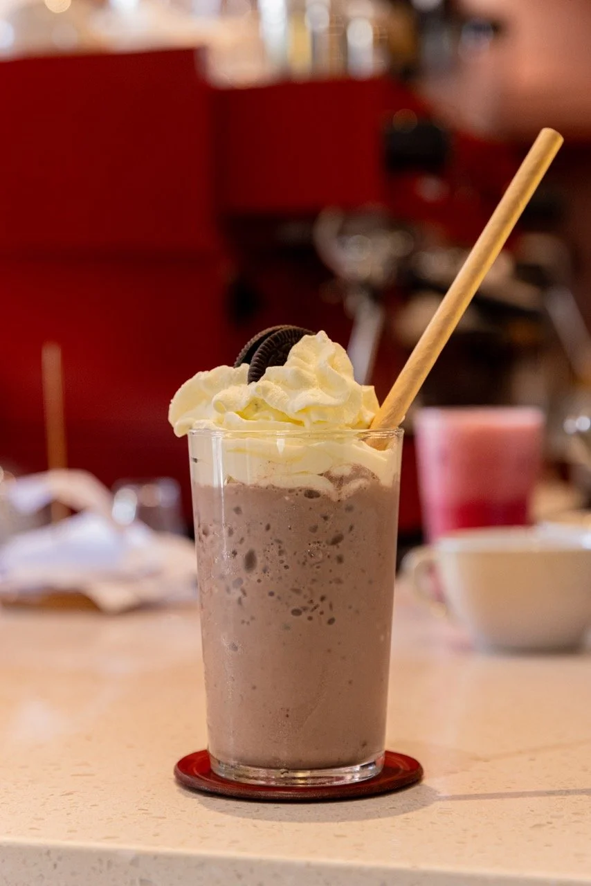 A tall glass of chocolate milkshake topped with whipped cream and an Oreo cookie, with a straw, on a bar counter.