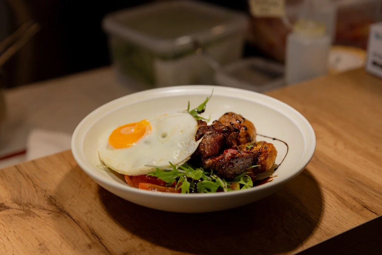 A white bowl with a fried egg, grilled meat, and fried plantains, garnished with greens and drizzled with sauce, placed on a wooden surface.