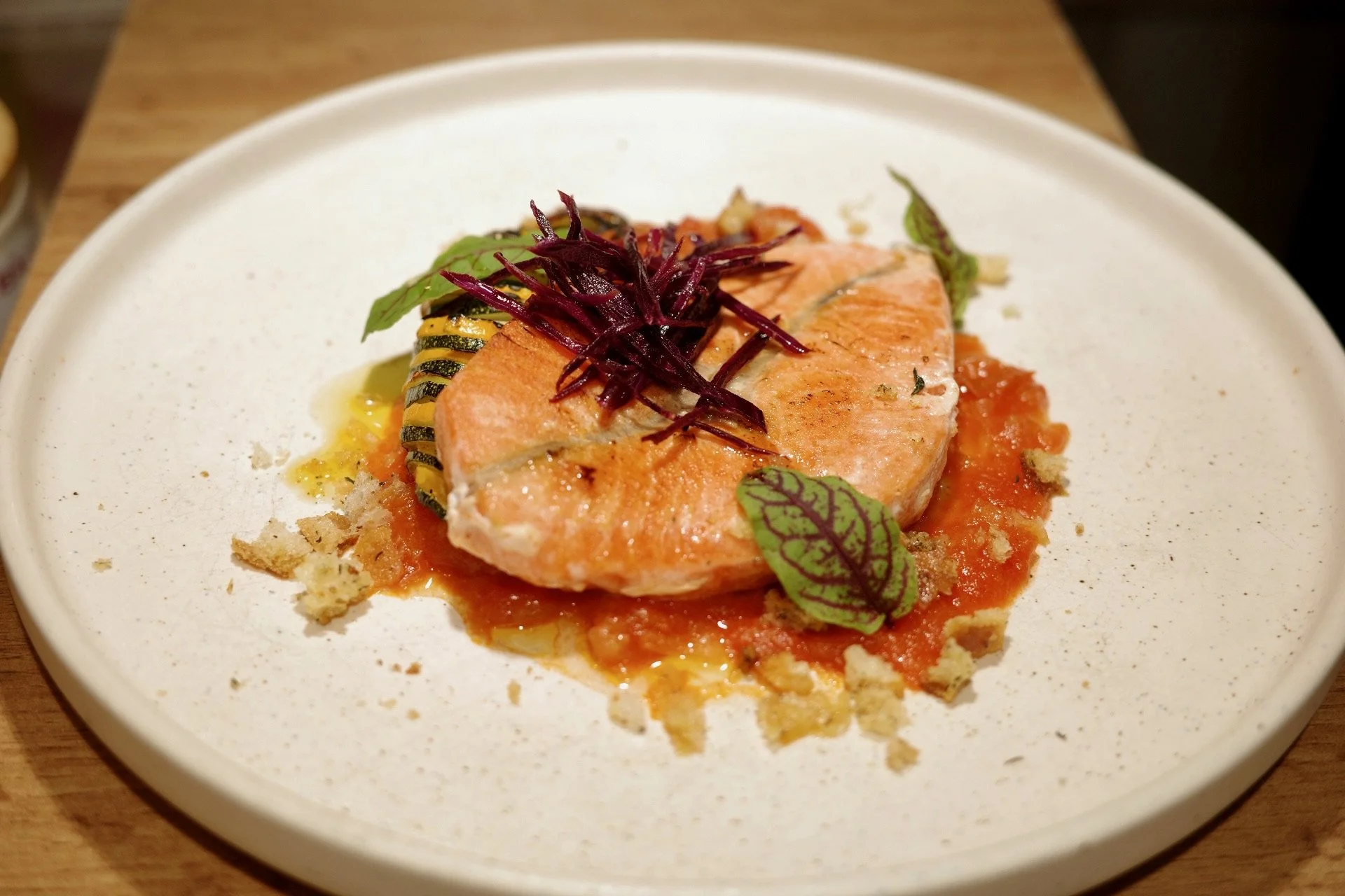 A plate with a cooked salmon fillet on top of tomato sauce, garnished with purple shredded vegetables and green herbs.