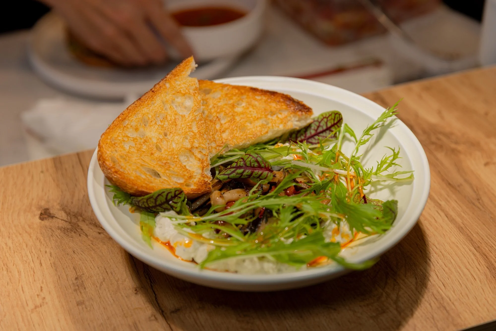 A white bowl contains a toasted bread slice and a mixed greens salad with various leafy greens, placed on a wooden surface.