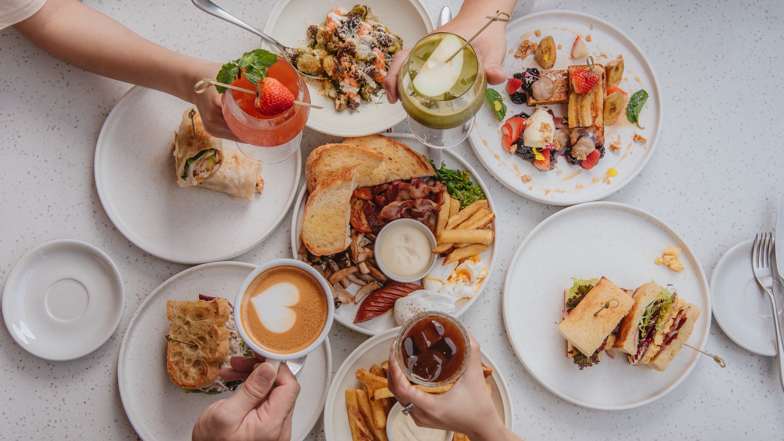 Top-down view of a table filled with various plates of food and drinks, including sandwiches, fries, a wrap, desserts, and beverages.