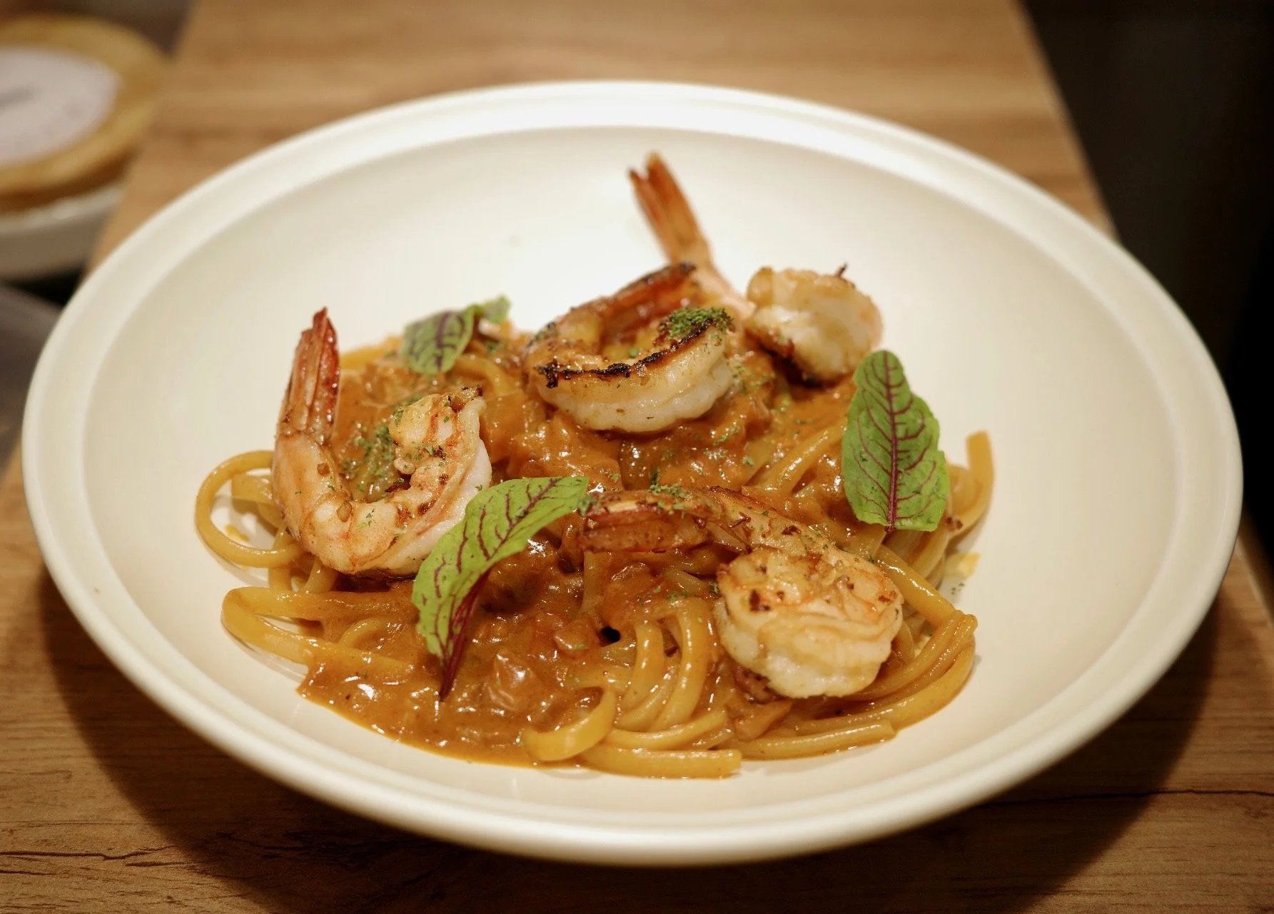 Bowl of shrimp pasta with sauce, garnished with small green leaves, served on a wooden table.