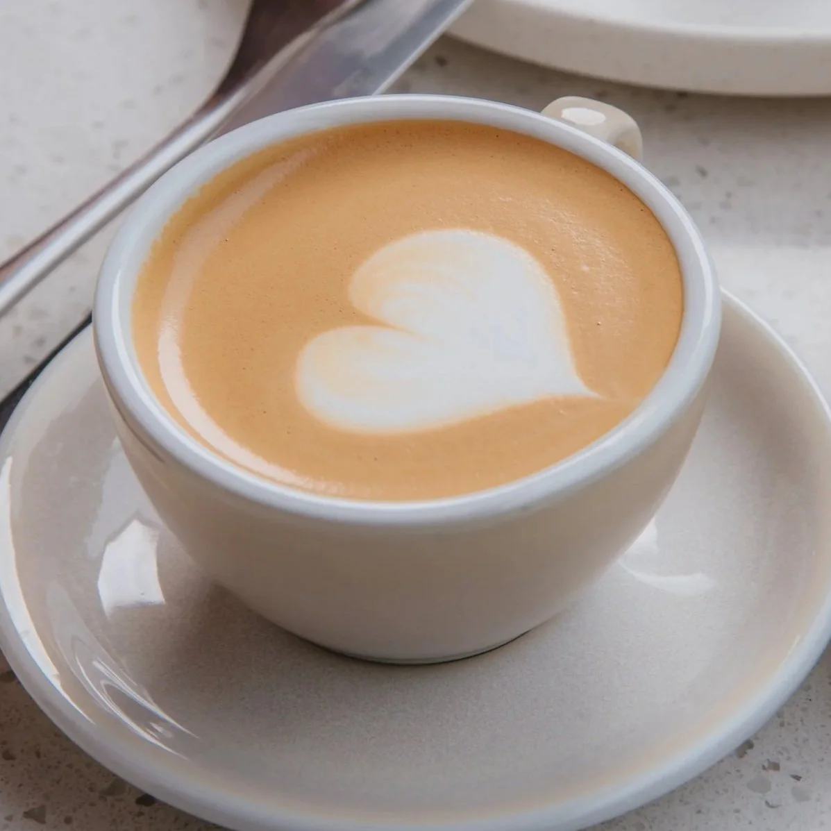 A white cup of coffee with foam art in a heart shape, sitting on a matching white saucer on a light-colored surface.