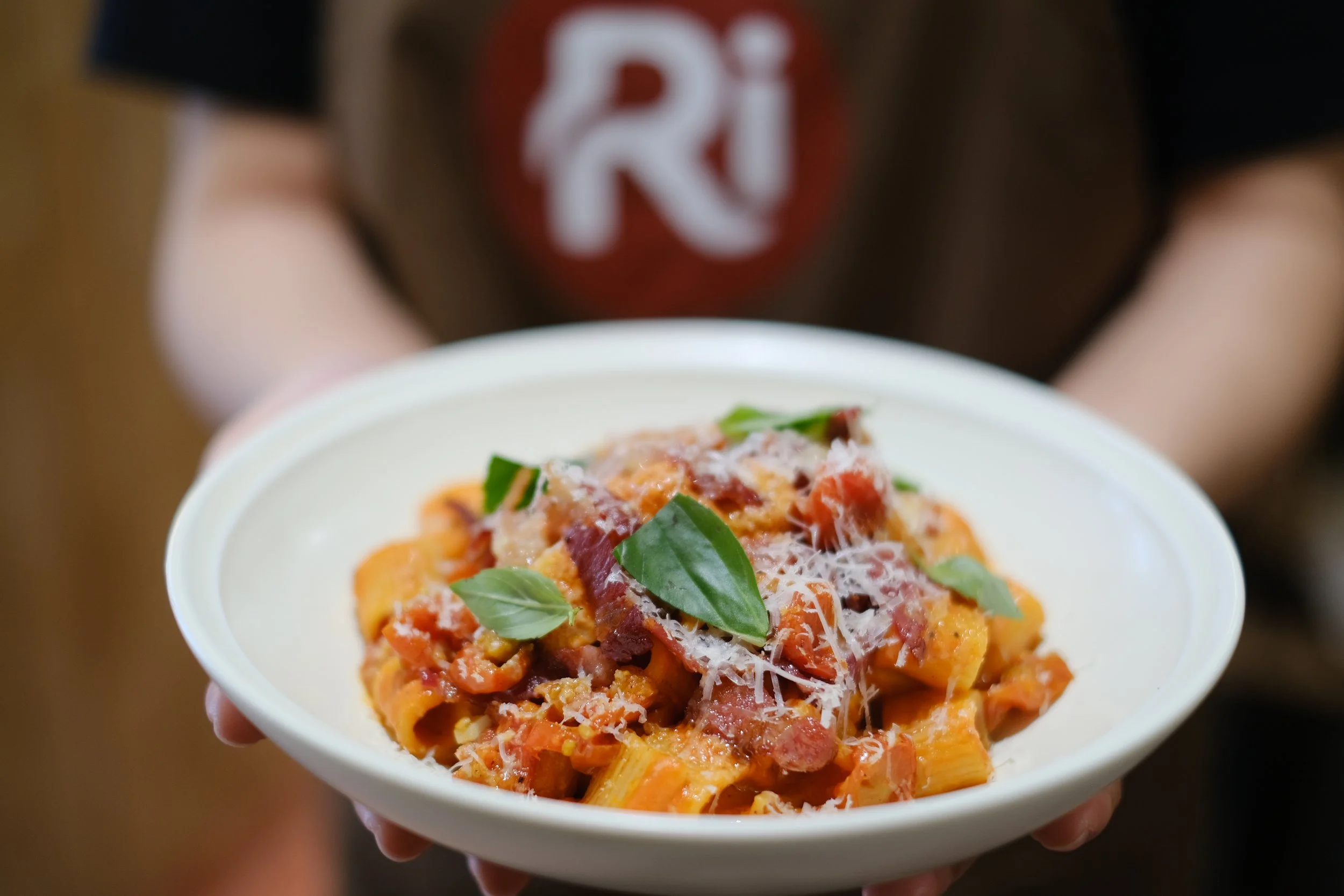 A person holding a white bowl filled with pasta, topped with chopped tomatoes, basil leaves, and grated cheese.