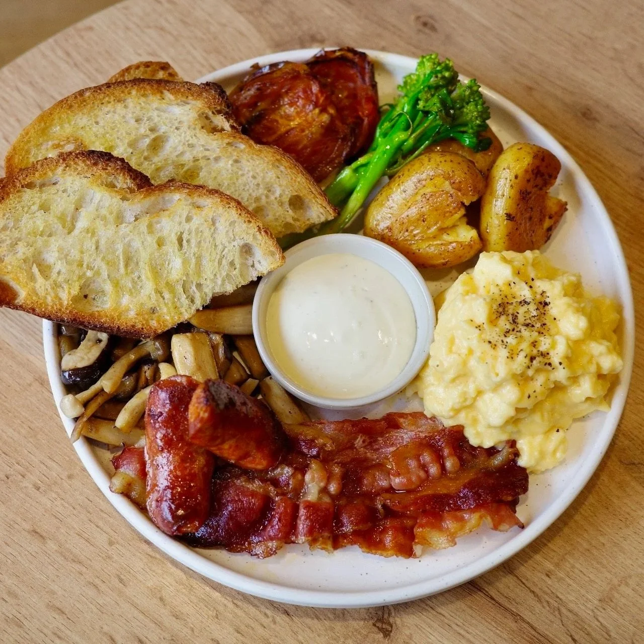 A plate of breakfast food including scrambled eggs, toast, sautéed mushrooms, bacon, roasted potatoes, broccoli, a tomato, chicken drumstick, and a small cup of white sauce.
