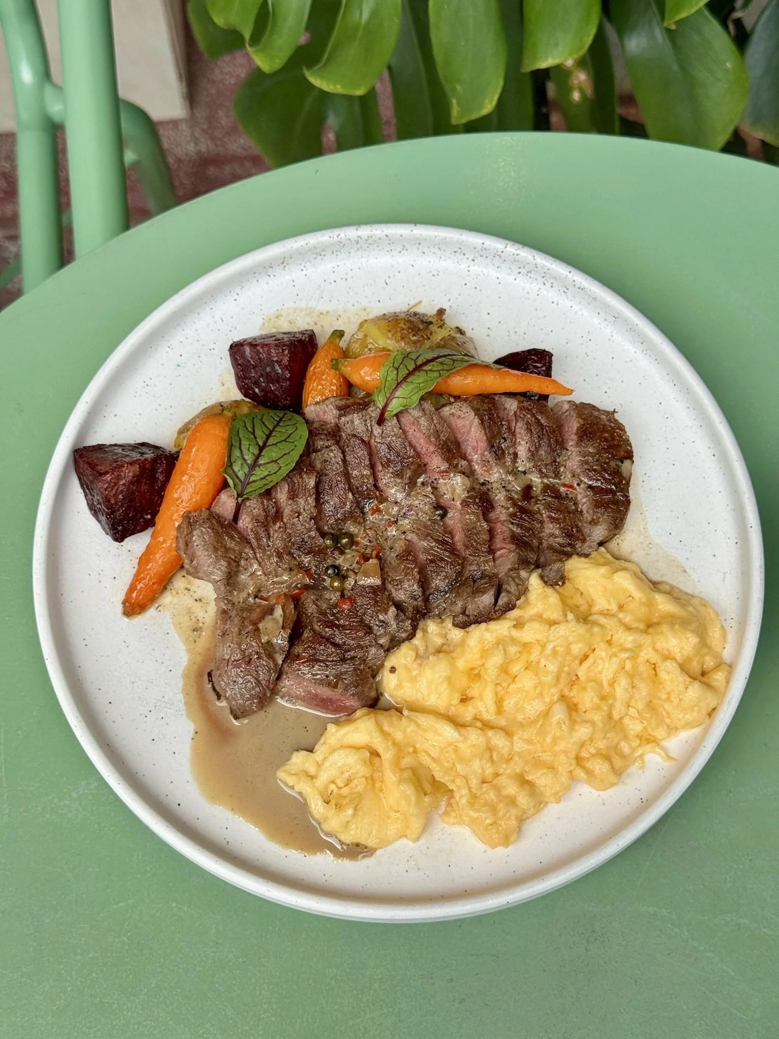 A plate of cooked food including mashed potatoes, sliced steak, roasted carrots, beets, and leafy herbs on top, on a green table.