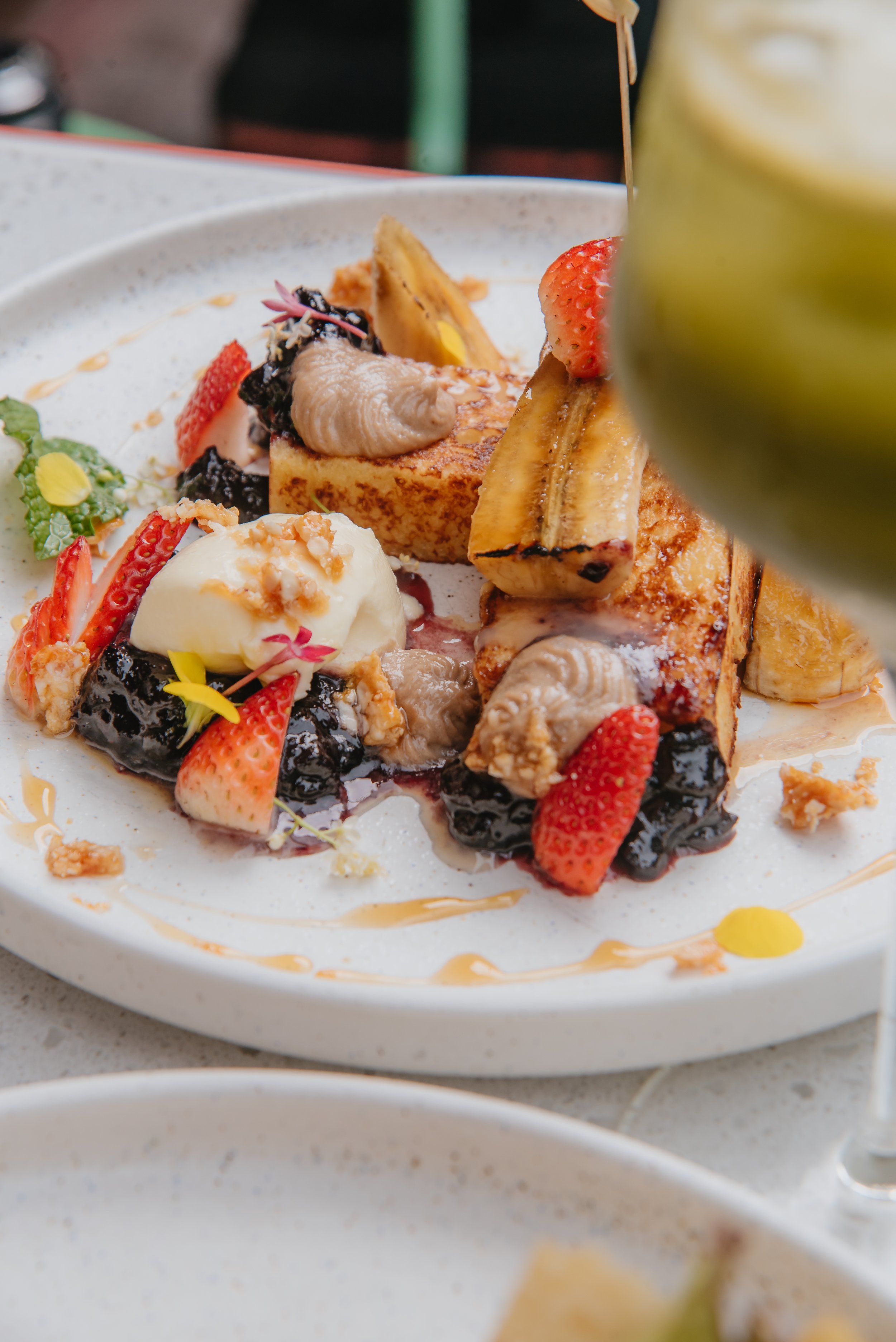 A plated dessert with grilled banana, strawberries, vanilla ice cream, and various toppings, garnished with edible flowers and syrup.