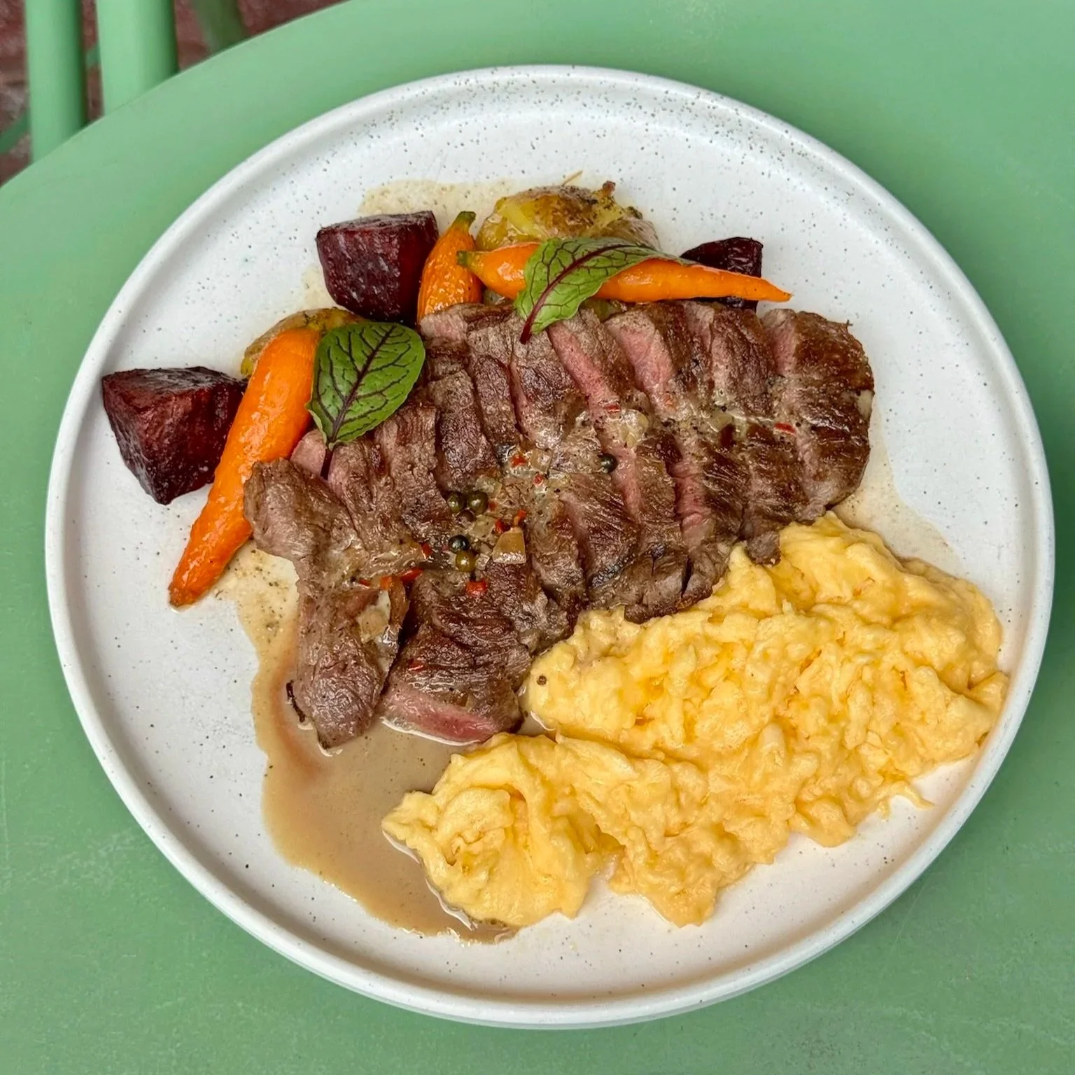 A plate of food featuring scrambled eggs, sliced steak with peppercorns, roasted carrots, beets, and green leaves.