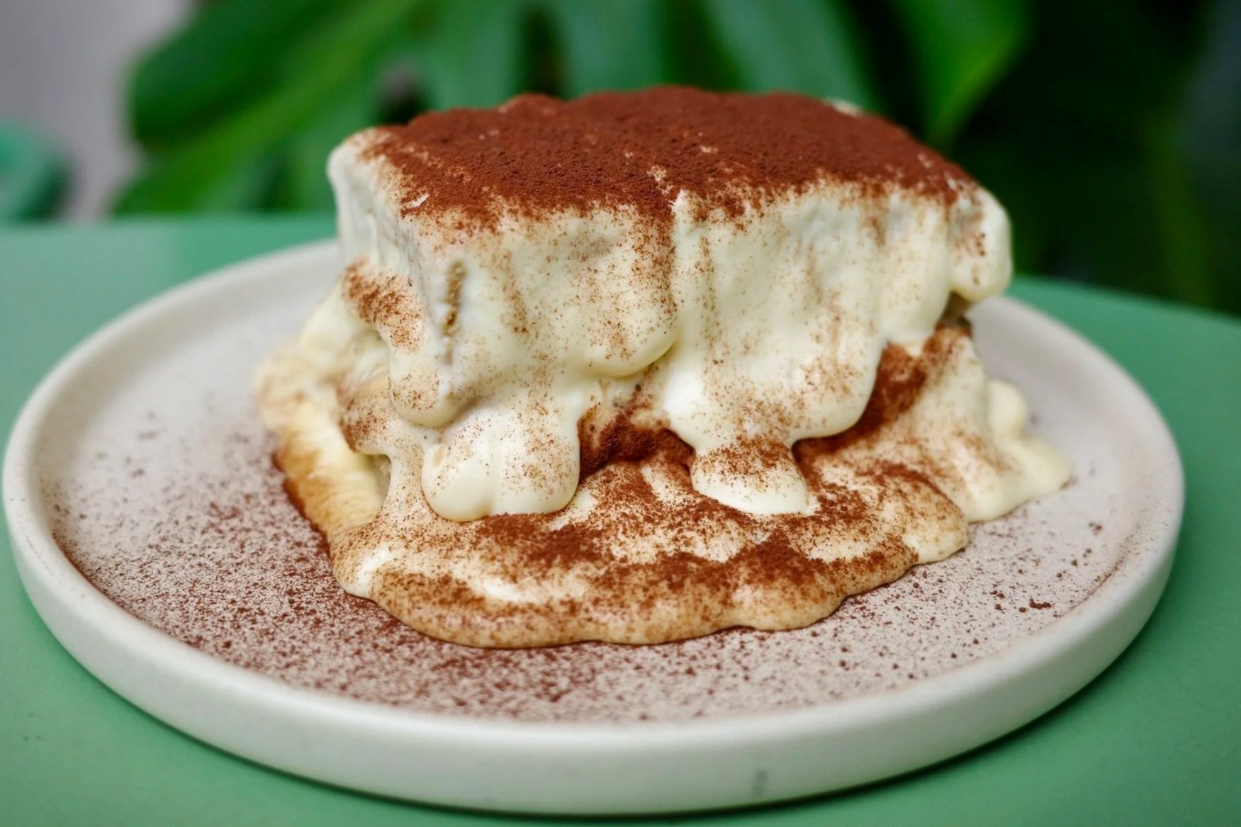 Close-up of a dessert with layers of cream, dusted with cocoa powder, in a white bowl on a green surface.