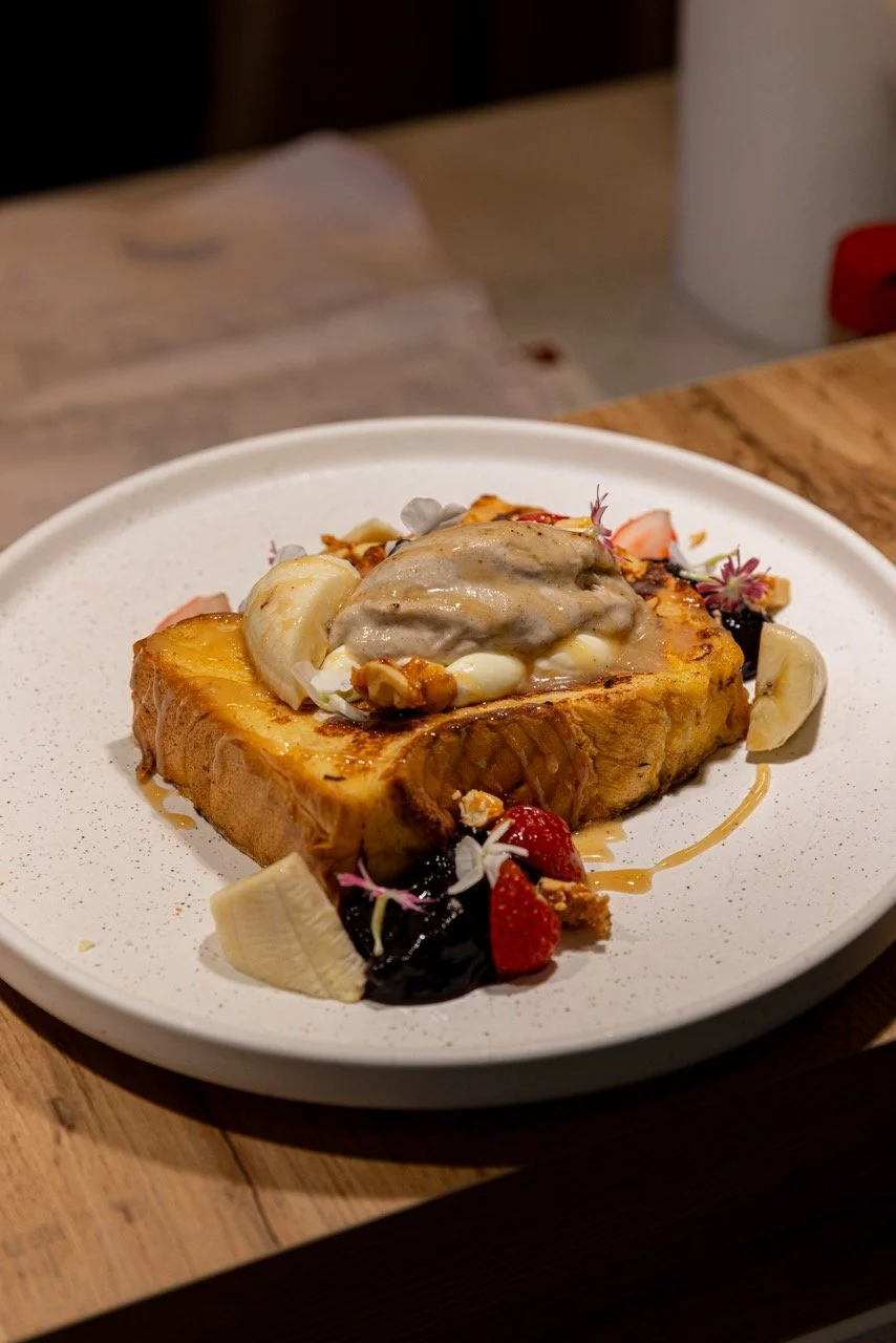 A plate of French toast topped with whipped cream, sliced bananas, strawberries, and edible flowers, drizzled with syrup.