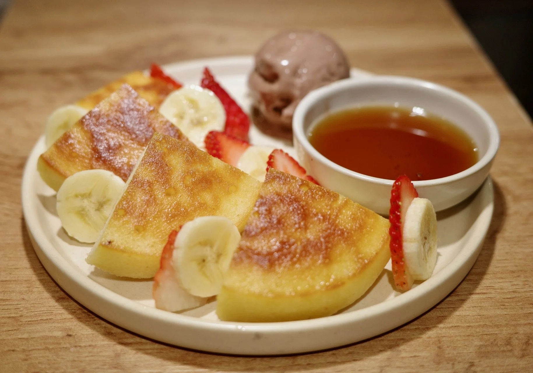 Plate with slices of French toast topped with sugar, banana slices, strawberry slices, a scoop of chocolate ice cream, a cup of tea, and a small bowl of syrup.