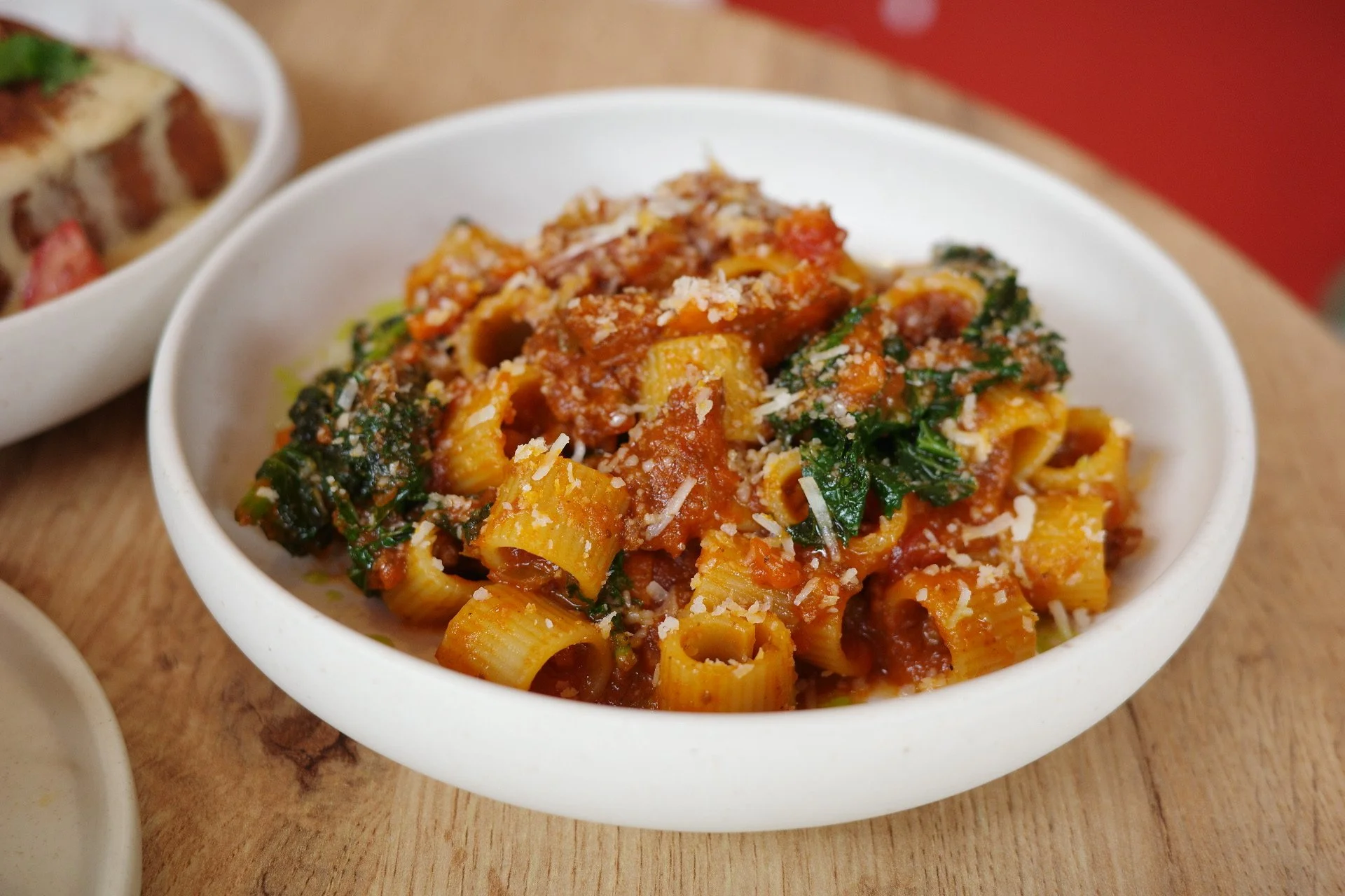 A bowl of cooked pasta with tomato sauce, spinach, grated cheese, and herbs on a wooden table.