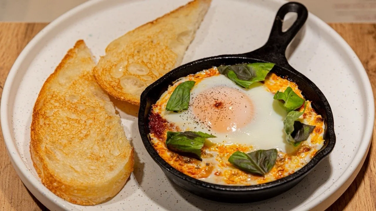 Fried eggs garnished with basil leaves served in a small cast iron skillet with toasted garlic bread slices on a white plate.