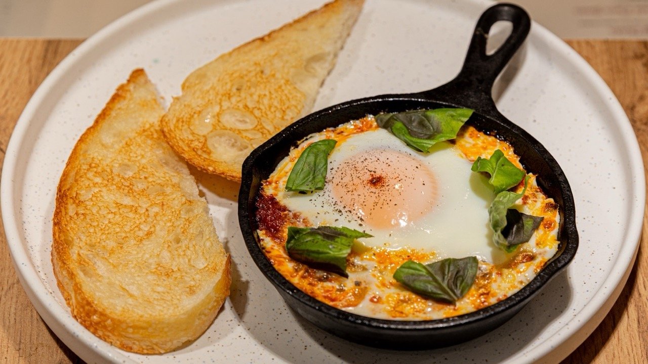 Fried egg with basil leaves in a small cast iron skillet, served with toasted bread slices on a speckled white plate.