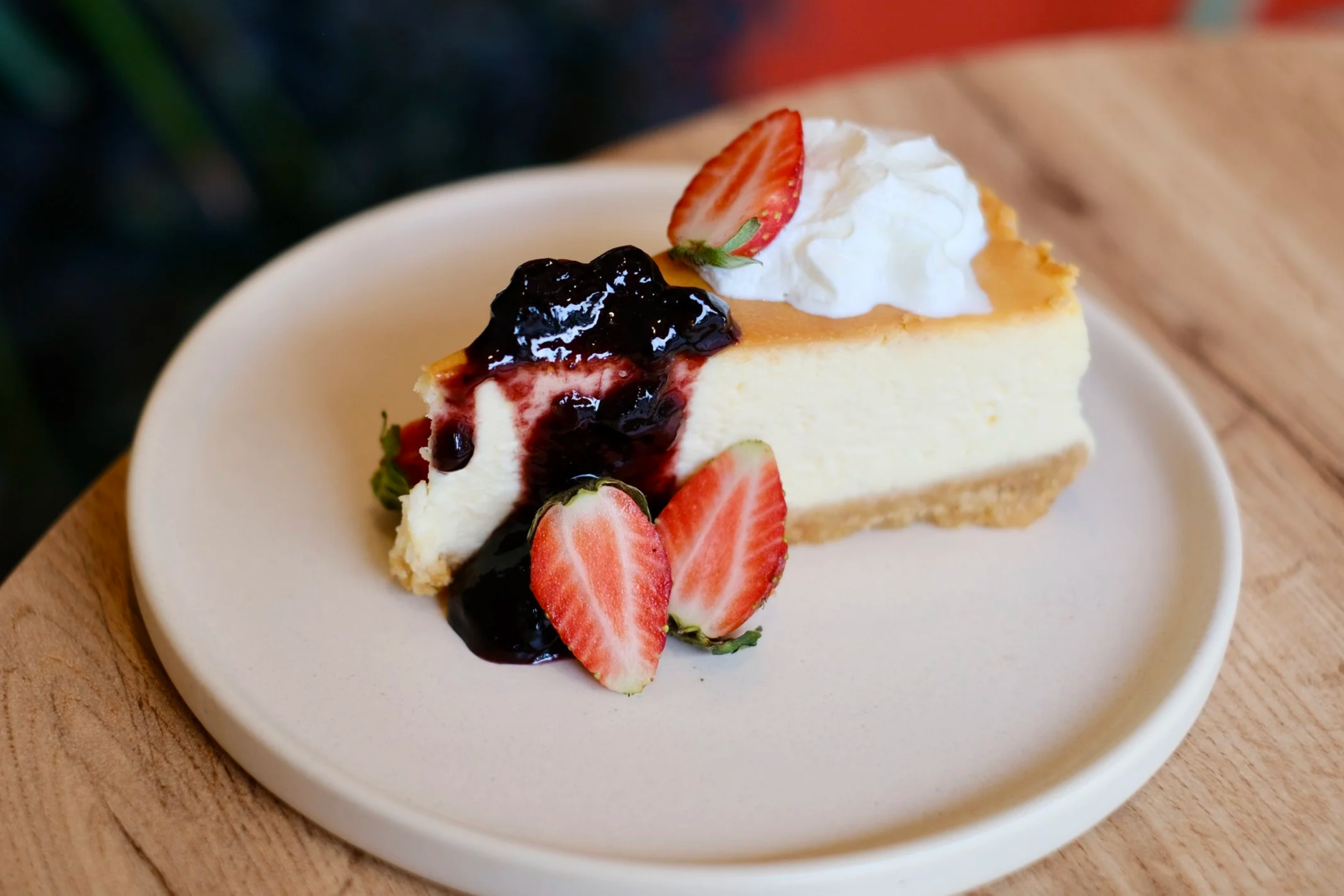 A slice of cheesecake topped with whipped cream, strawberry slices, and blueberry compote on a white plate and wooden surface.