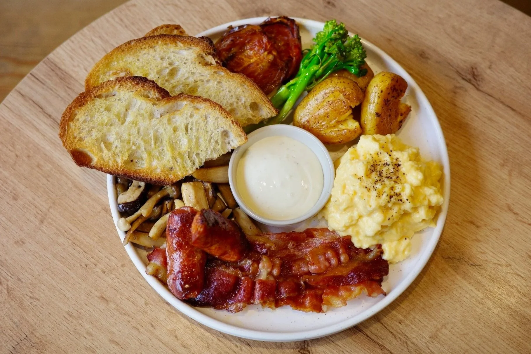 A white plate with toast, roasted potatoes, broccoli, mushrooms, sausage, bacon, scrambled eggs, meatball, and dipping sauce on a wooden table.