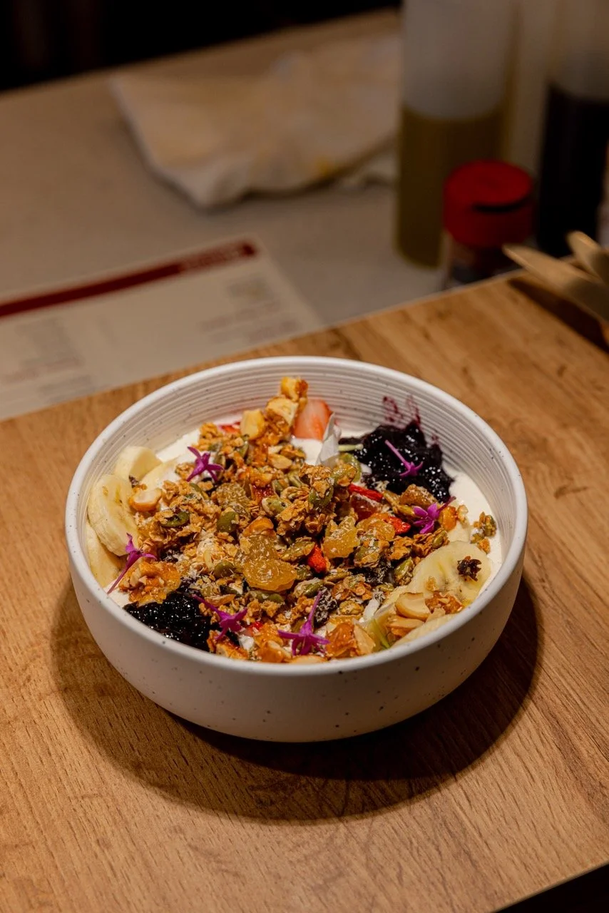 Bowl of yogurt topped with granola, banana slices, berries, and edible flowers on a wooden table.