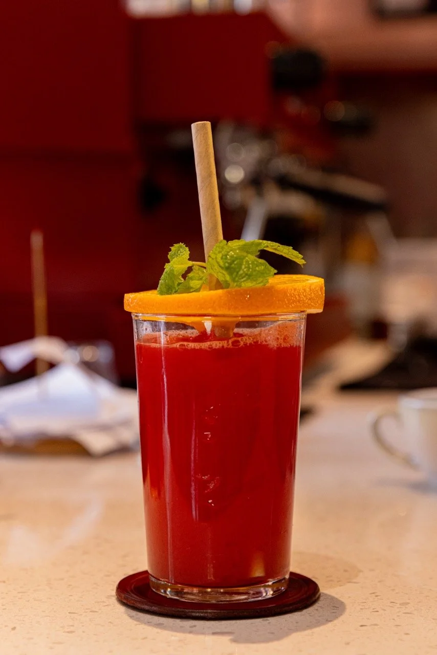 A tall glass of red cocktail garnished with an orange slice and a sprig of mint, with a straw on a coaster, on a light-colored table.