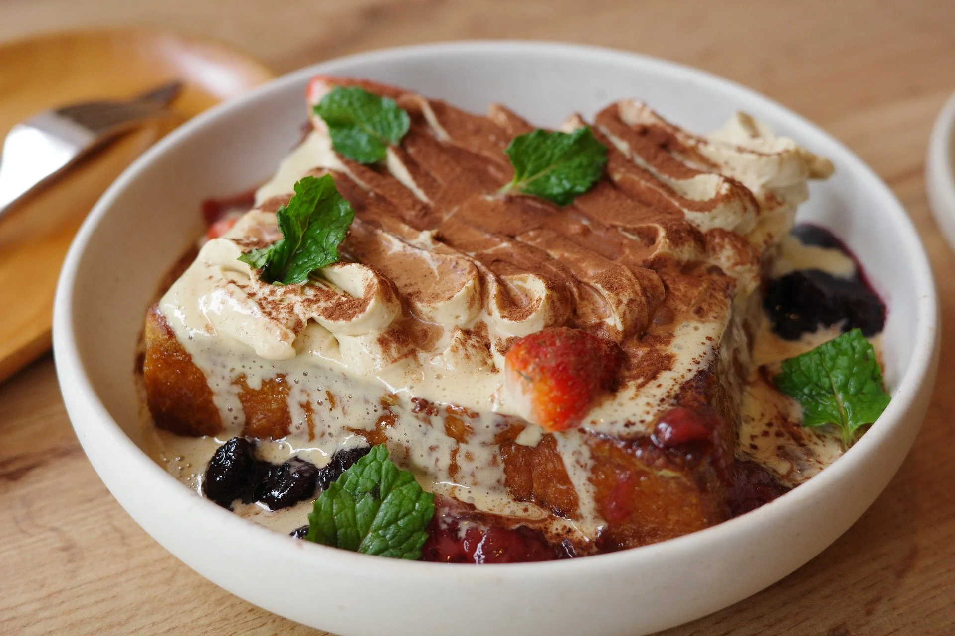Dessert parfait with whipped cream, cocoa powder, mint leaves, fresh strawberries, blueberries, and blackberries in a white bowl.