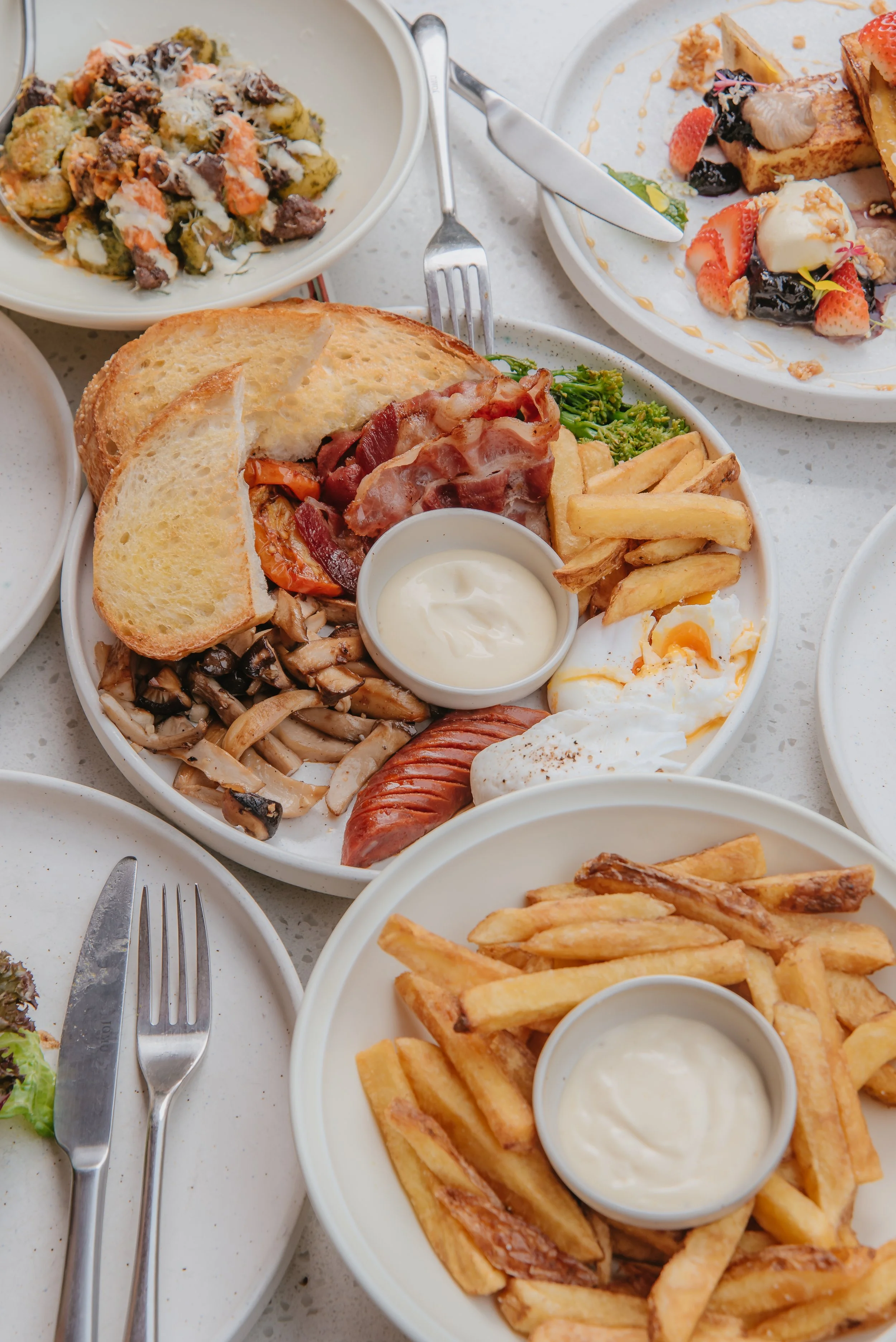 A breakfast plate with toast, bacon, sausage, scrambled eggs, French fries, sautéed mushrooms, and a small cup of white sauce, surrounded by other breakfast dishes including pasta and waffle desserts.