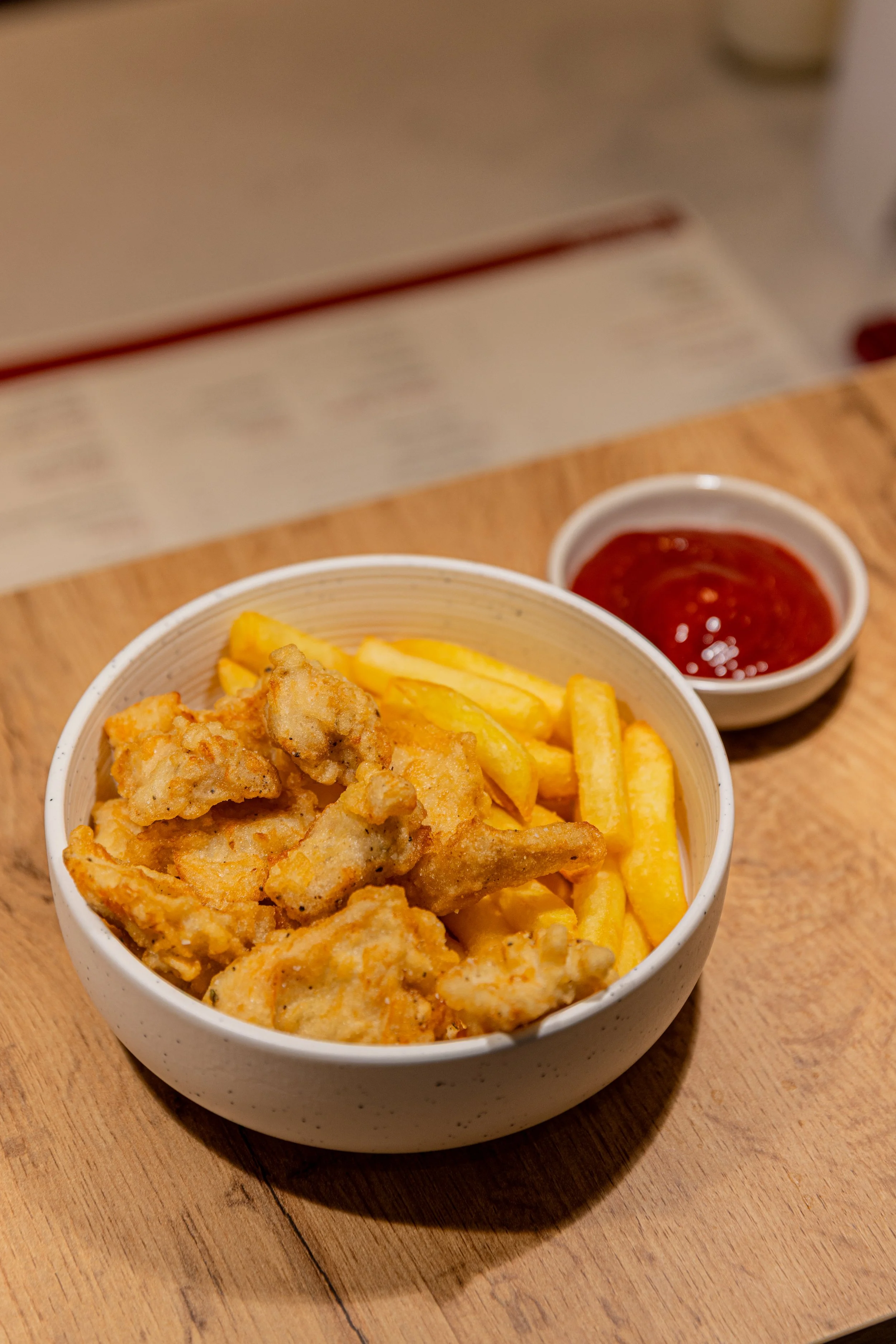 Close-up of a bowl containing fried chicken pieces, French fries, and a small container of ketchup, on a wooden table.