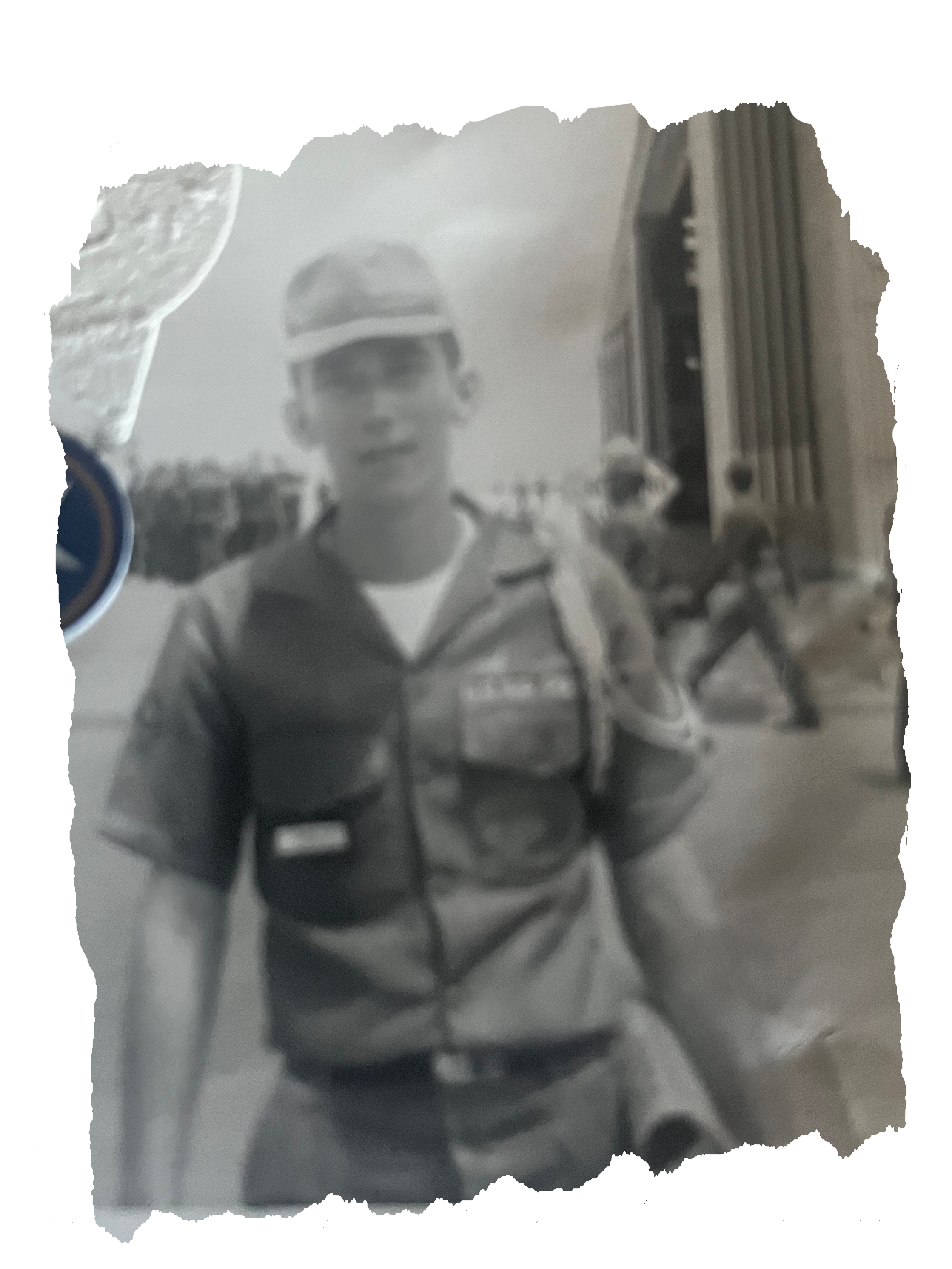 Black and white photo of a young man wearing a cap and sunglasses, standing outdoors with buildings and people in the background. Vietnam Veteran Papa