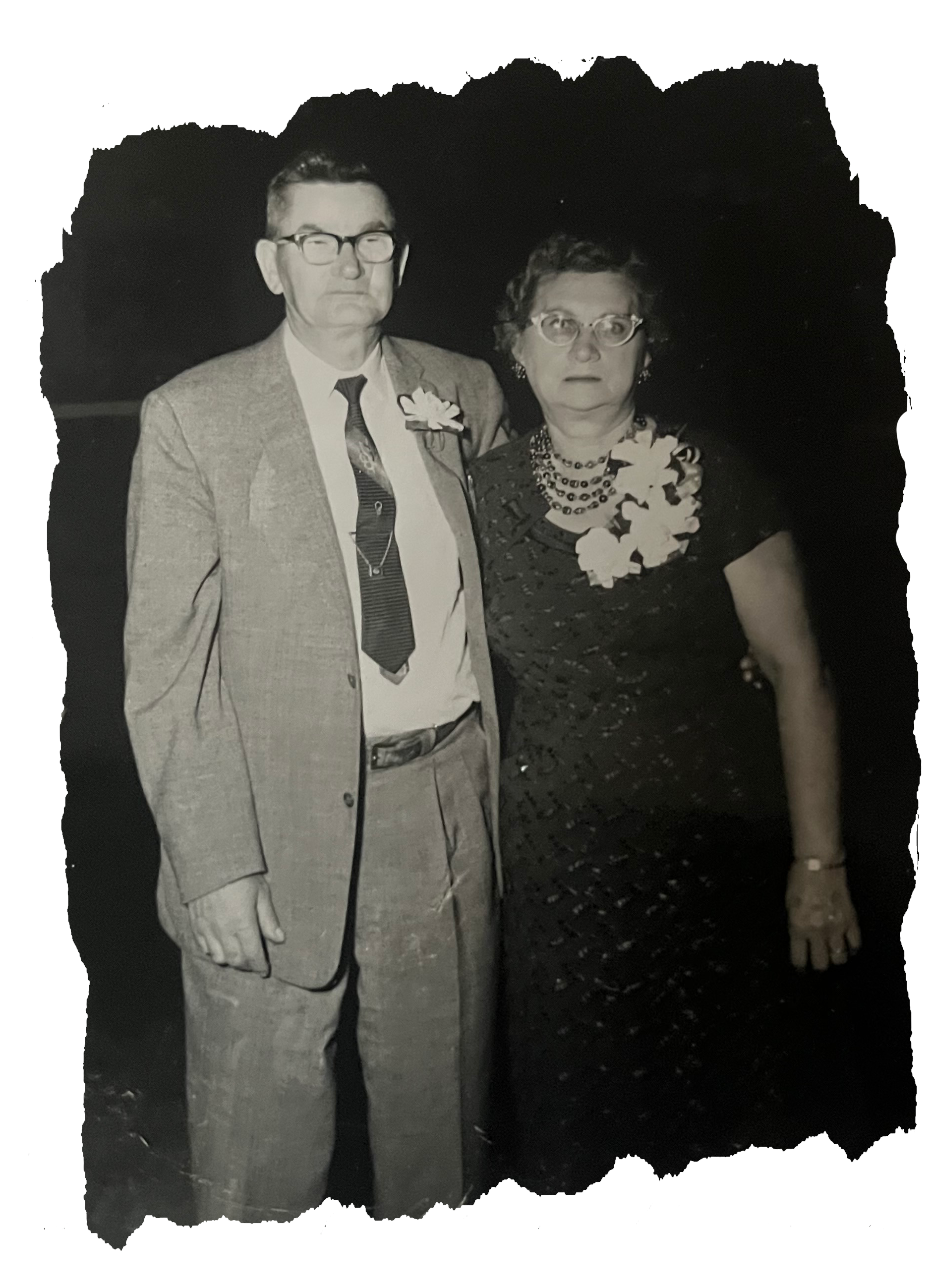 Black and white photo of a man and woman standing together inFormal attire, both wearing floral corsages, in a dimly lit setting.  Babcia and Dziadek