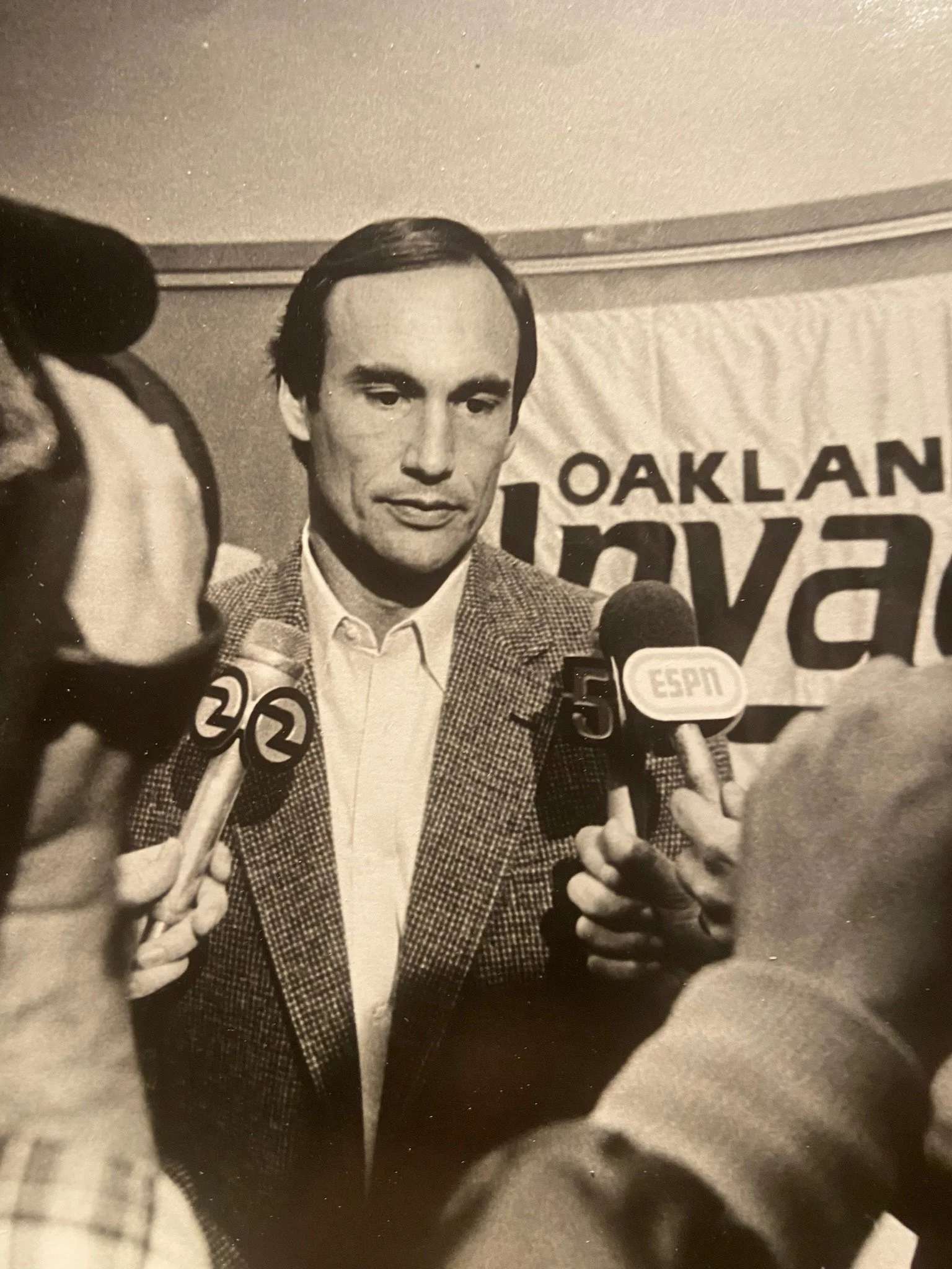 Black and white photo of a man speaking to reporters, wearing a suit jacket, collared shirt, and tie. Several microphones with news station logos are in front of him, and a banner with partially visible text is in the background.