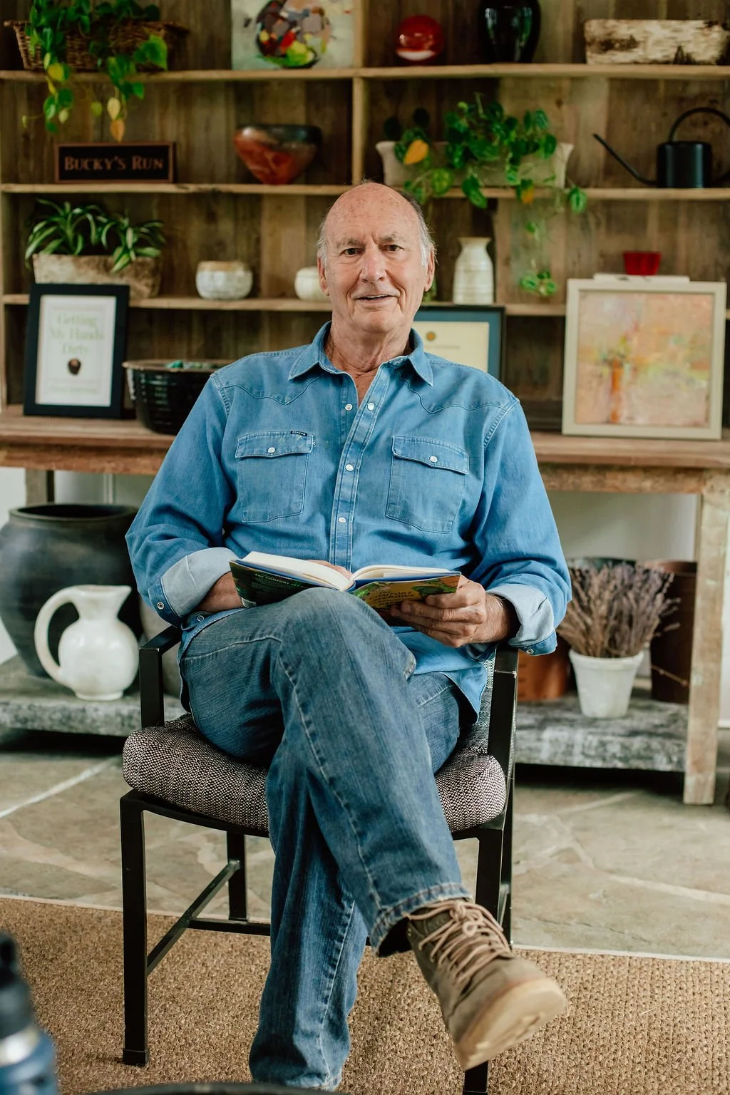 An elderly man with a bald head and light skin, wearing a blue denim shirt and jeans, sits on a chair reading a book in a cozy indoor setting with wooden shelves filled with plants, vases, and framed art behind him.
