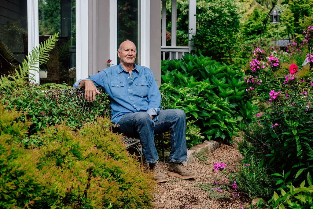 Older man sitting on a bench in a garden surrounded by green plants and purple flowers, near a house with large windows.