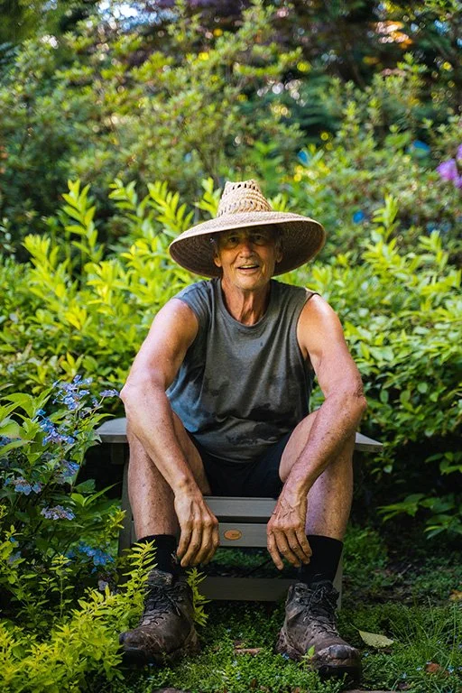 Author Chuck Hutchison in gardening clothes and hat sitting on a chair in his garden