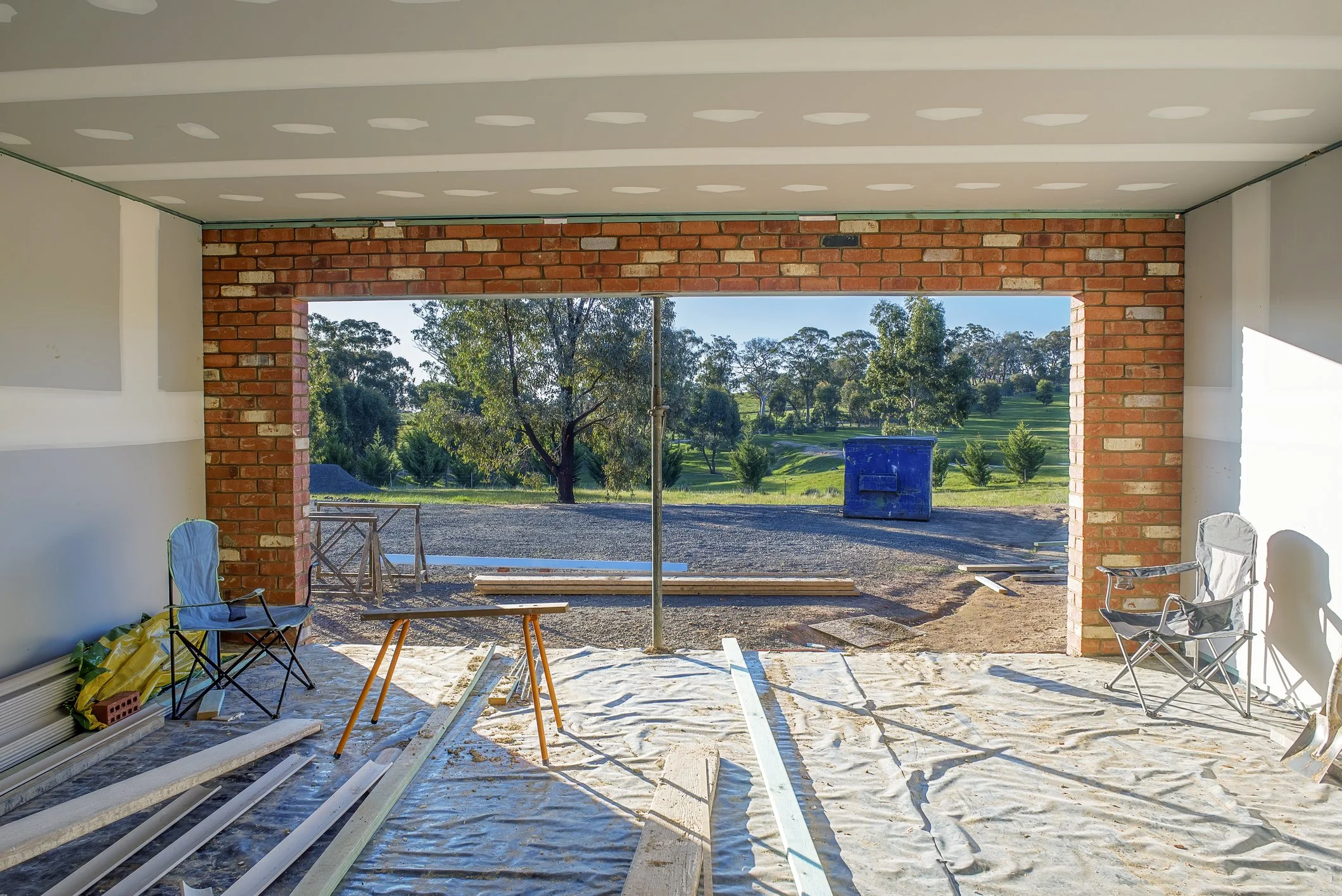 View from inside a house under construction, looking out through a large open wall with brick framing, showing a grassy yard with trees and a blue trash bin outside.