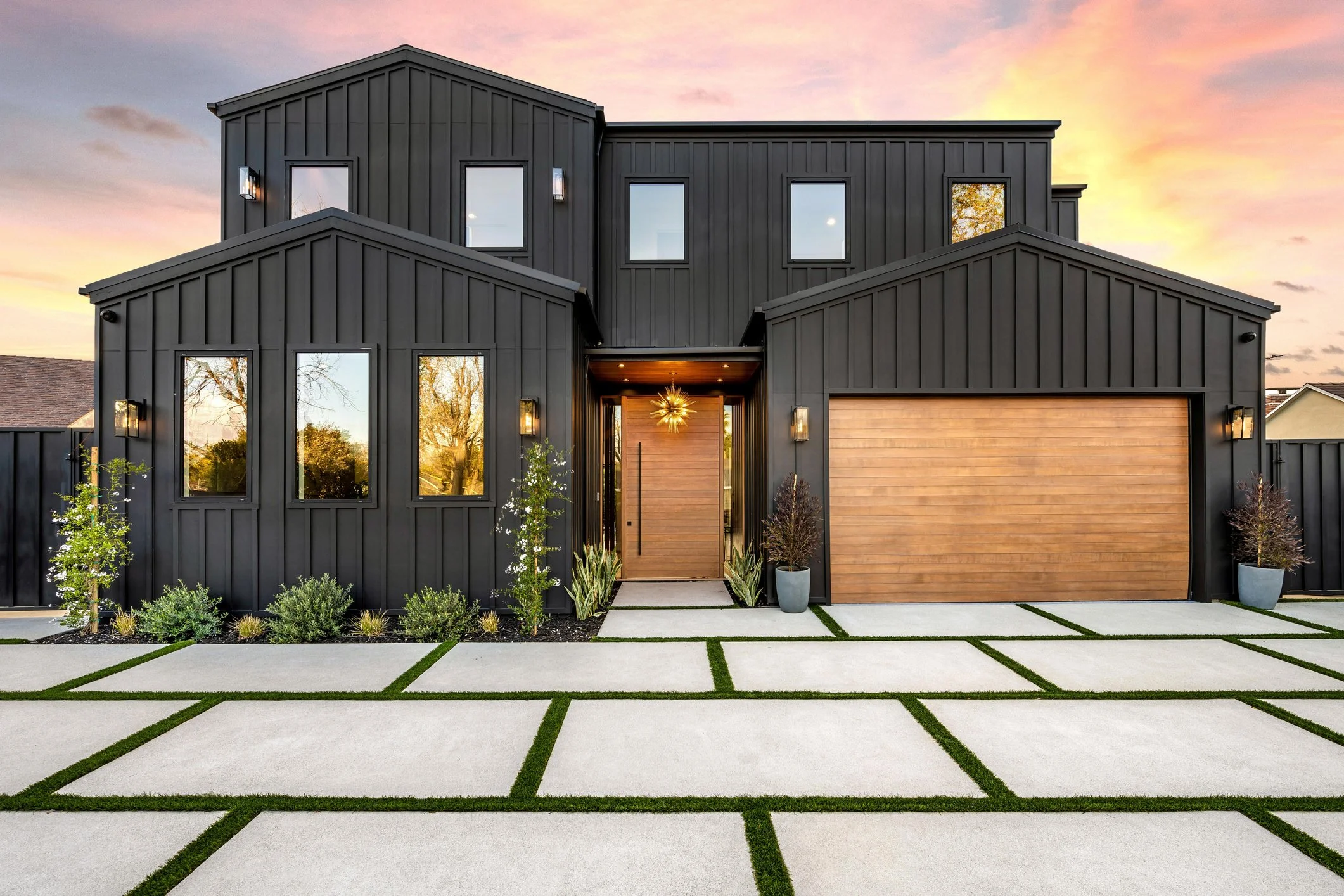 Modern two-story house with black exterior siding, wooden garage door, and front door, landscaped front yard with concrete pavers and green grass, trees and plants, sunset sky in the background.