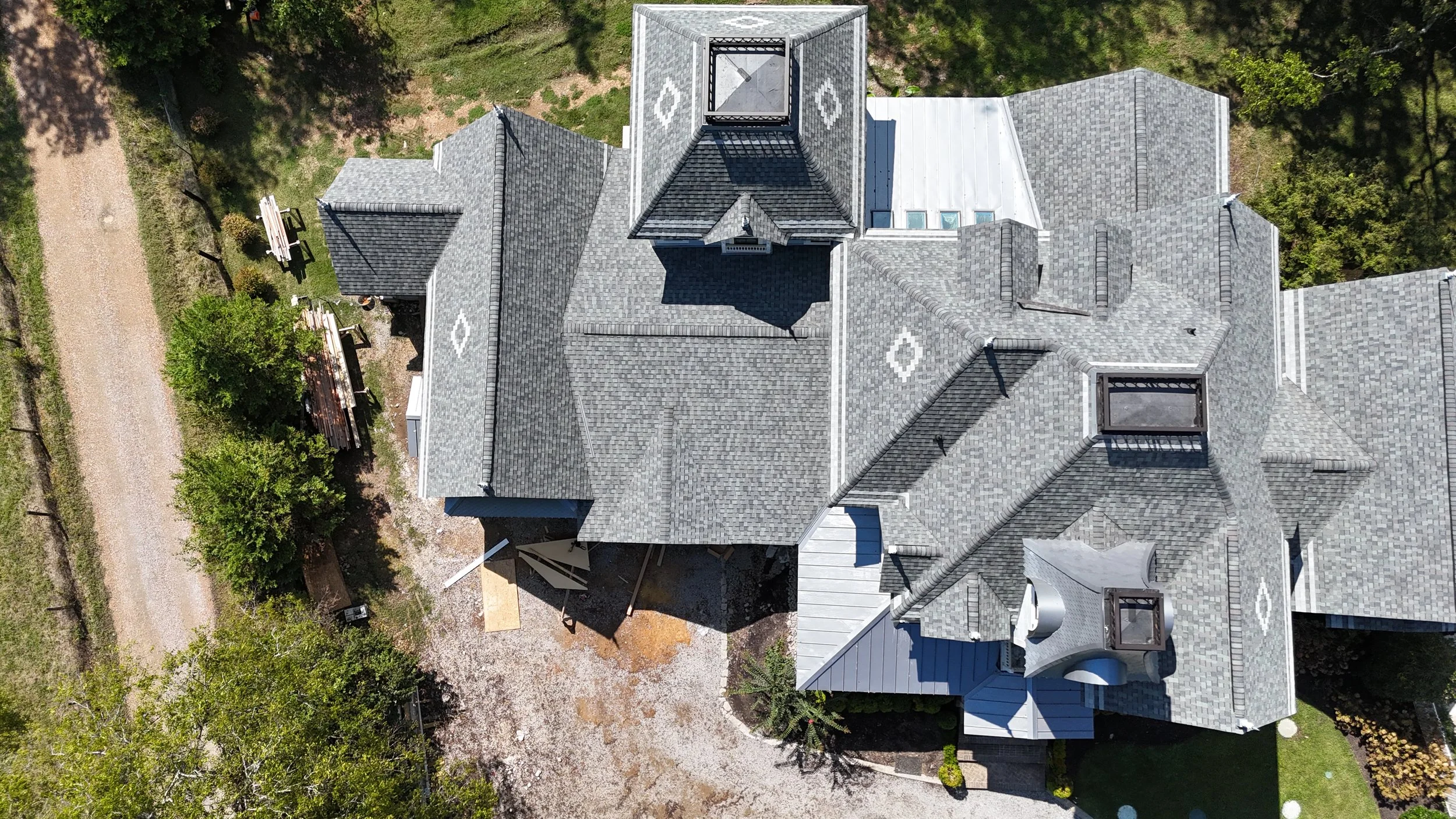 An aerial view of a house with gray shingle roof, multiple gables, dormer windows, and surrounding greenery and trees.