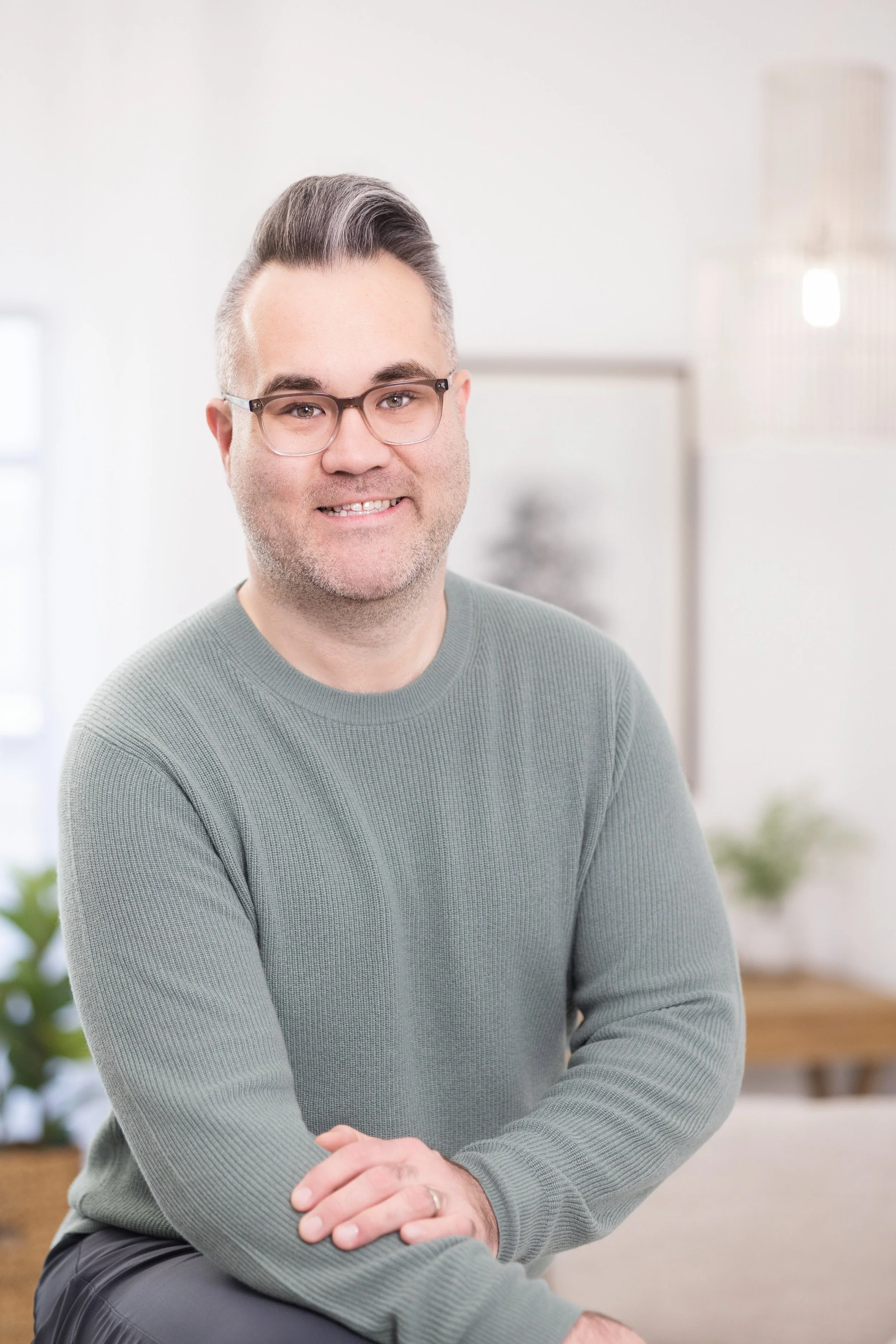 A man with glasses and gray hair smiling, sitting in a bright, modern room with minimal decor and plants in the background.