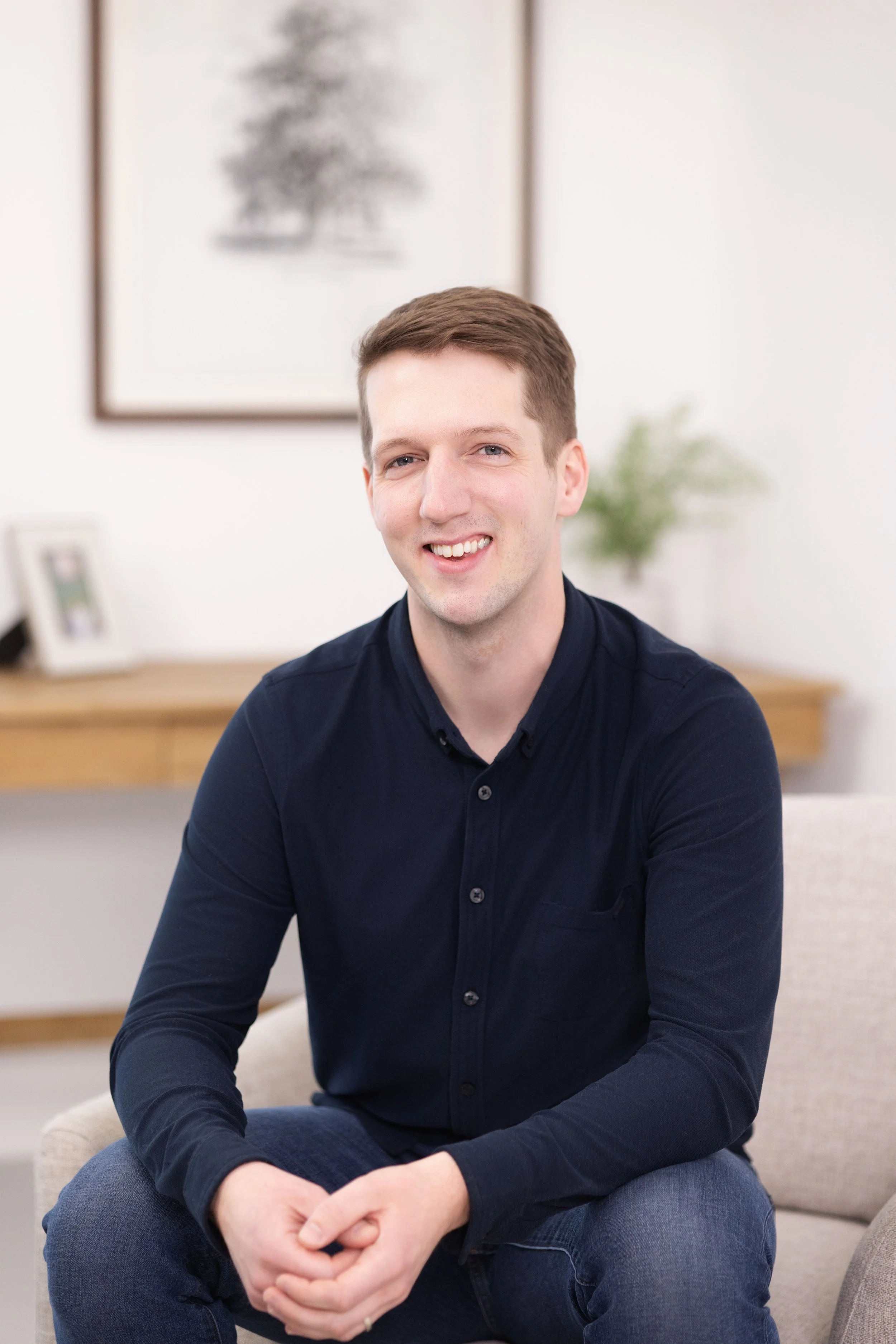 A young man with short brown hair and blue eyes sitting on a beige armchair, smiling at the camera in a bright room with framed artwork and a small potted plant in the background.