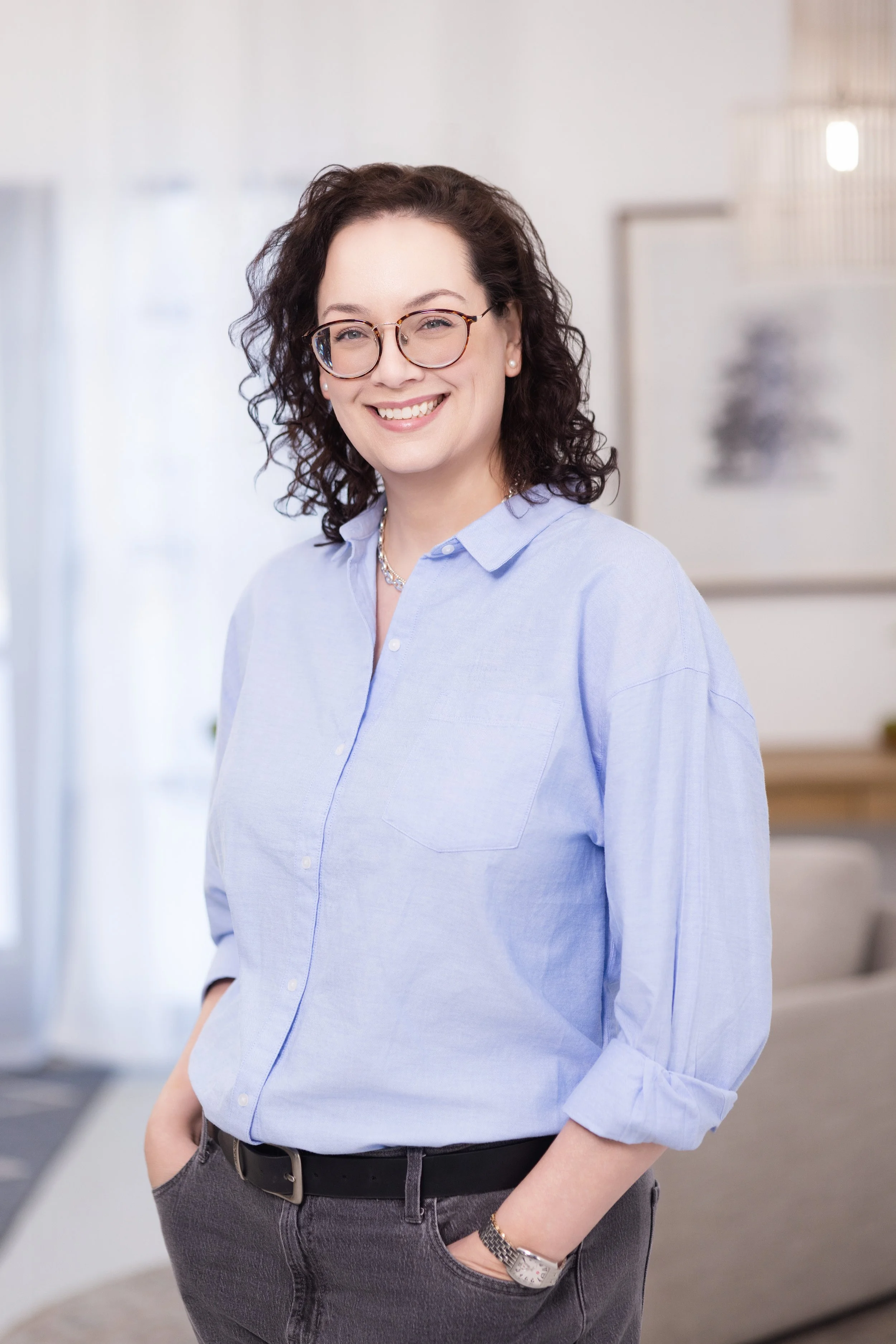 A woman with curly dark hair, glasses, and a light blue button-up shirt smiling indoors.