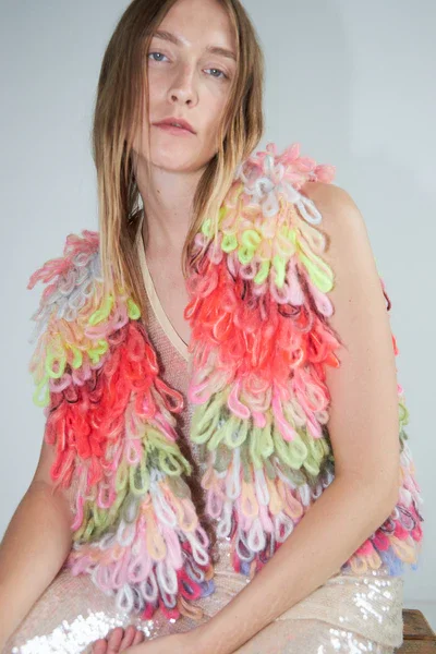 Frau in bunter, pelzähnlicher Weste mit Pastellfarben, sitzend auf einem Stuhl vor weißem Hintergrund.