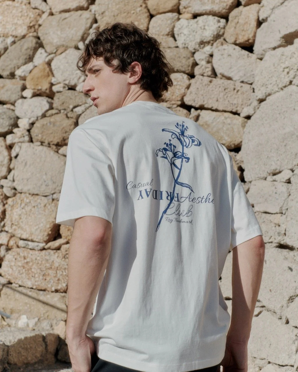 Junge trägt weißes T-Shirt mit blauer Blumenzeichnung und Text, steht vor einem Steinelement im Freien.