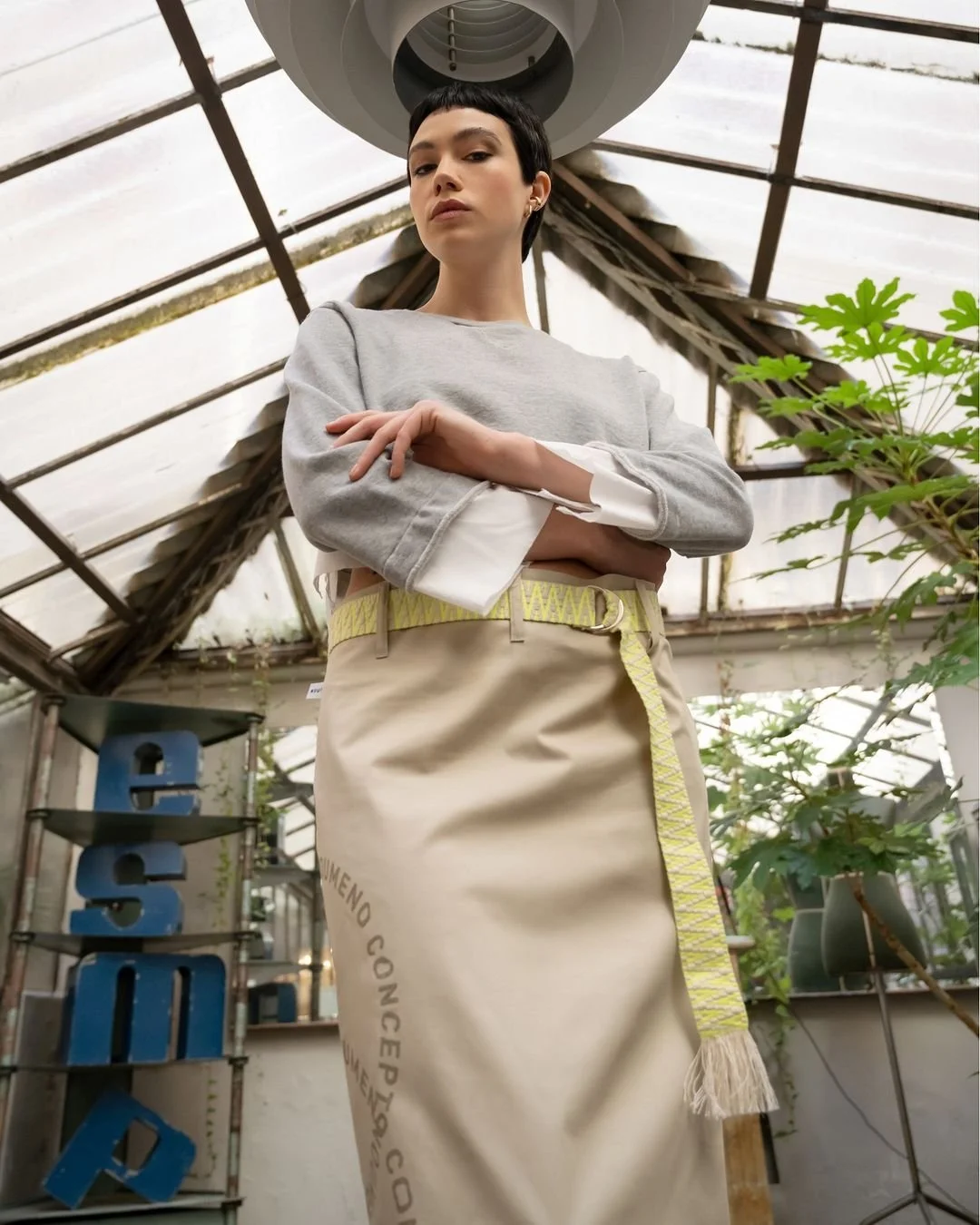 Junge Frau in einem Gewächshaus, trägt graues Sweatshirt und beige Rock mit gelbem Gürtel, blickt ernst in die Kamera.