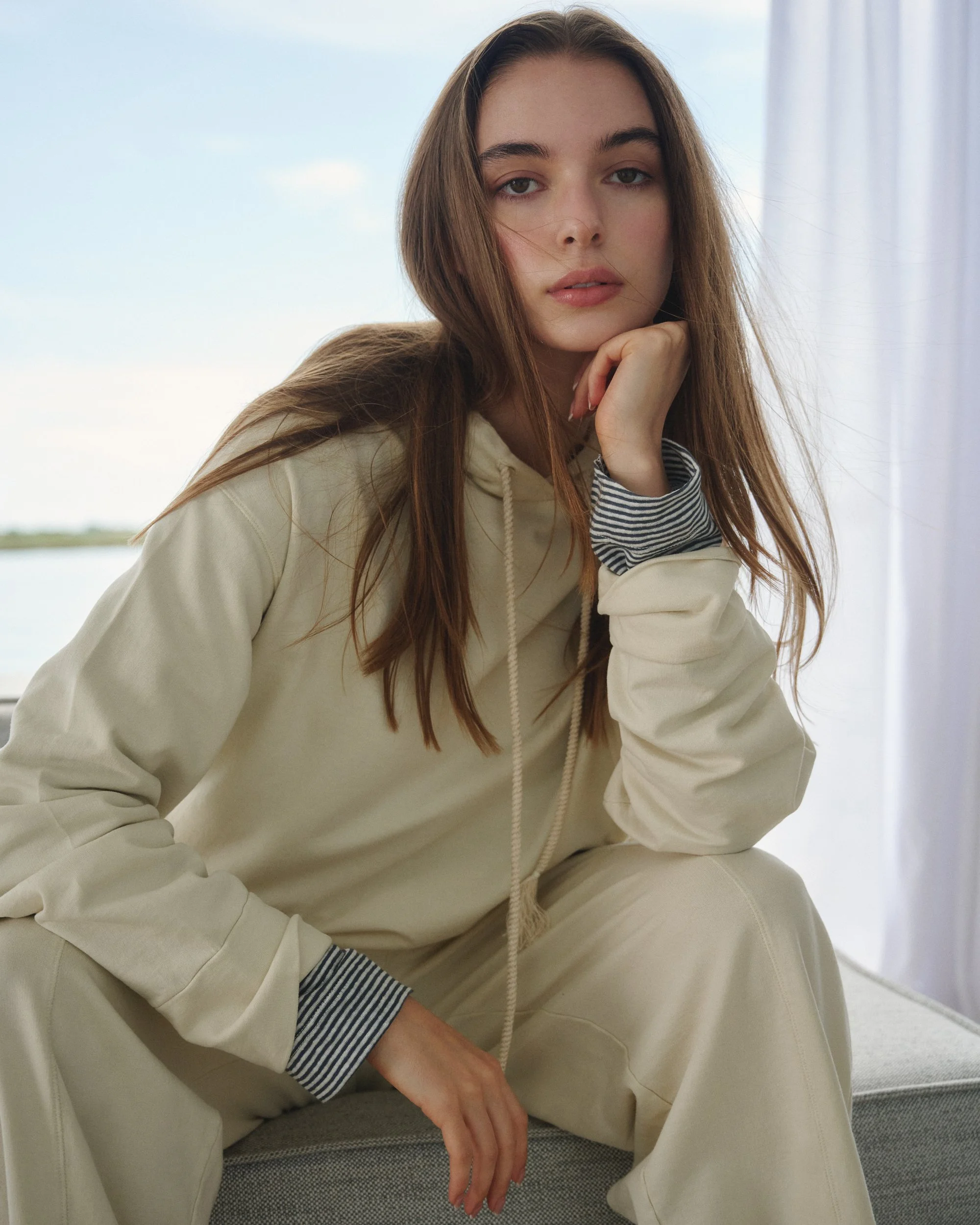 Junge Frau in bequemer Freizeitkleidung sitzt auf einem Sofa vor einem Fenster mit Sicht auf einen Fluss im Hintergrund.