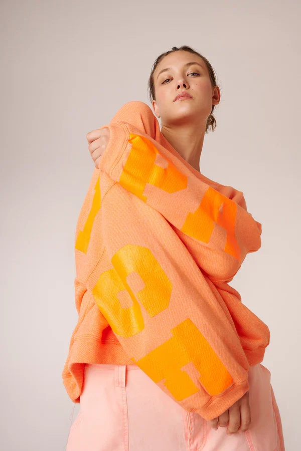 Frau trägt einen orangefarbenen Pullover mit großen gelben Buchstabenmuster und hellrosa Hosen.