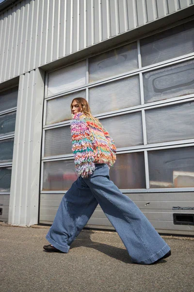 Frau in buntem Strickpullover und weiten Jeans posiert vor einer riesigen industriellen Garage