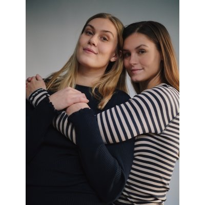 Zwei junge Frauen umarmen sich freundlich und schauen in die Kamera.
